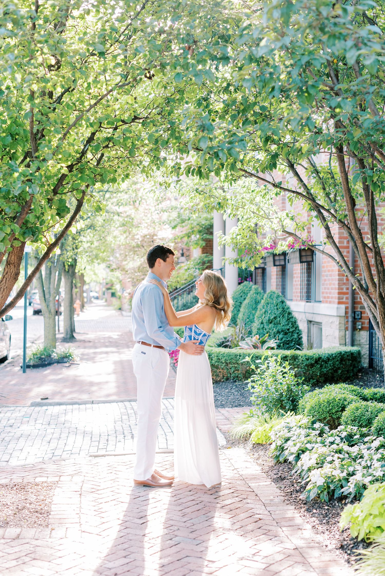 A romantic couple shares tender moments in a garden walkway lined with lush green trees and brick pavers.