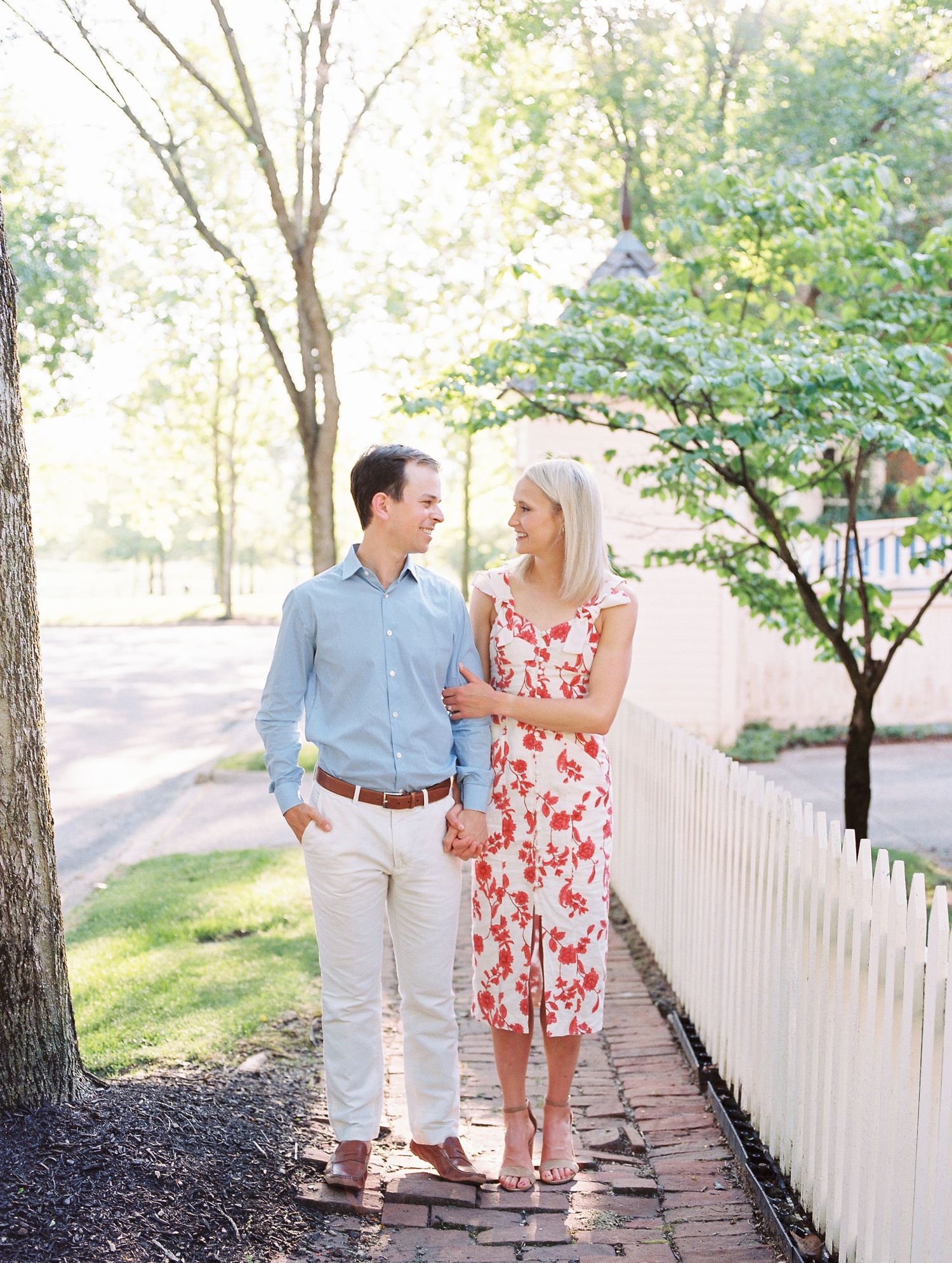 A couple strolls together on a brick path beside a white picket fence on a sunny spring day.