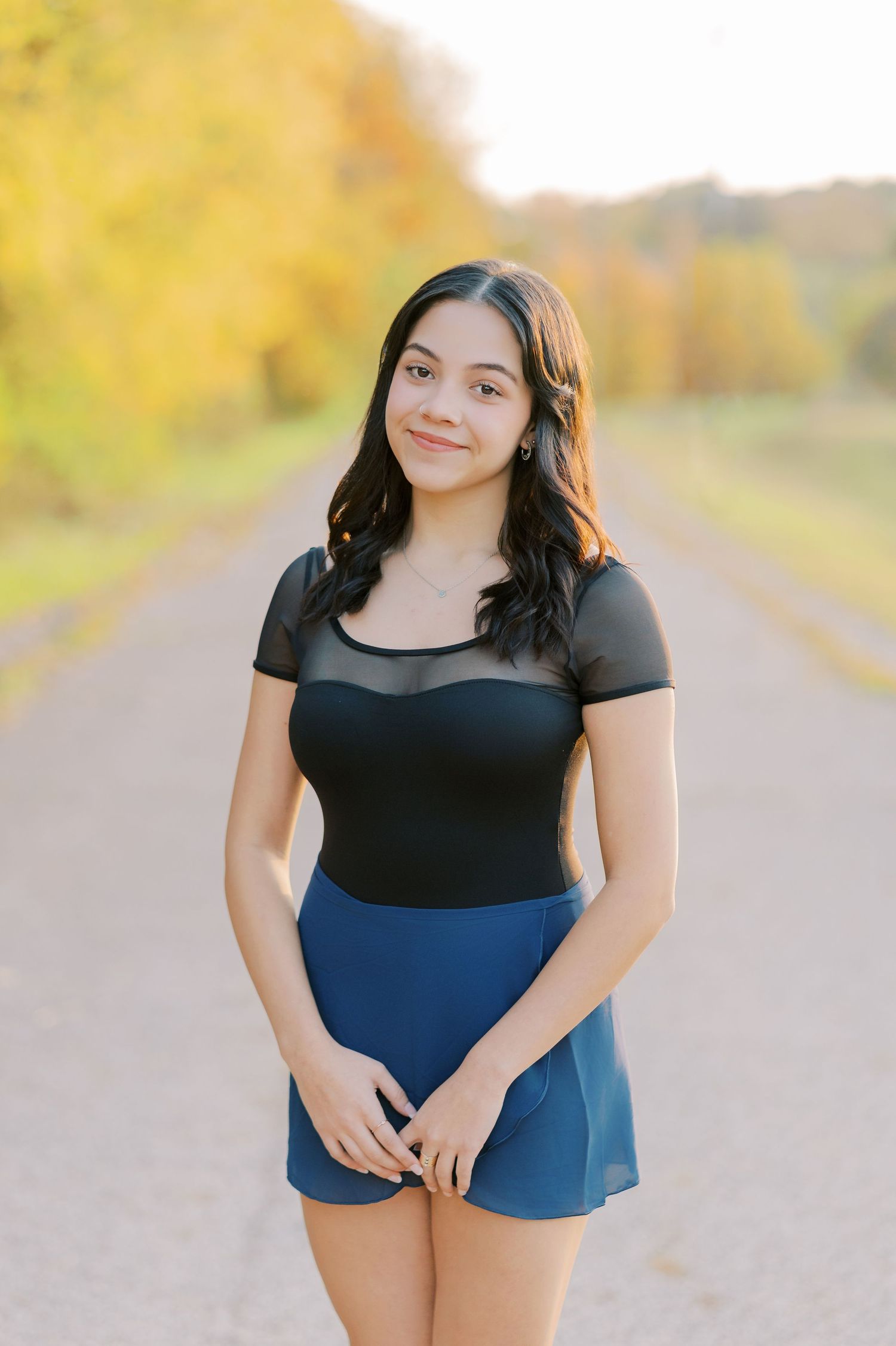 A person in a black and navy blue dress poses outdoors on a path during autumn.