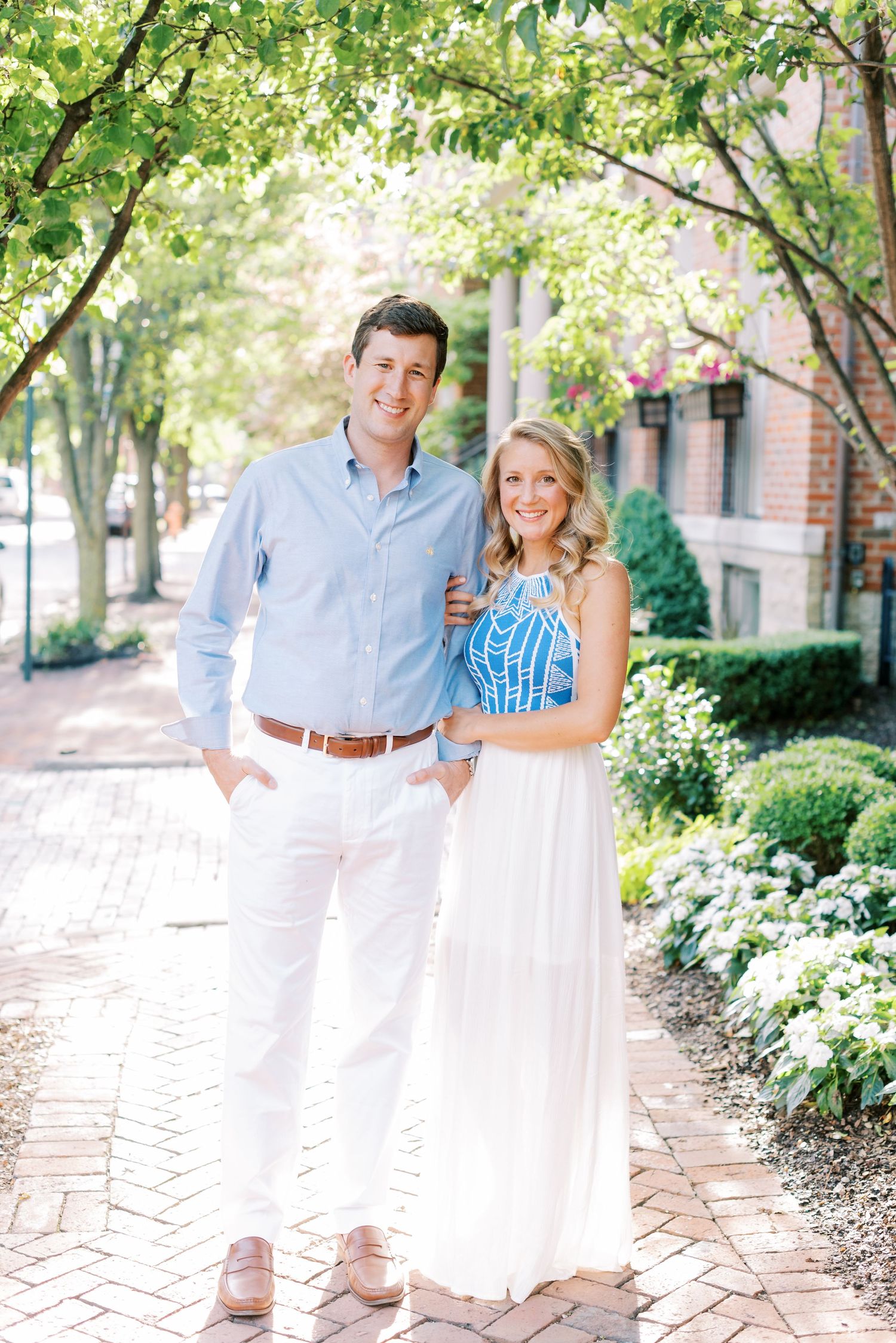 A couple in coordinated white and blue outfits poses together on a brick walkway surrounded by lush greenery.