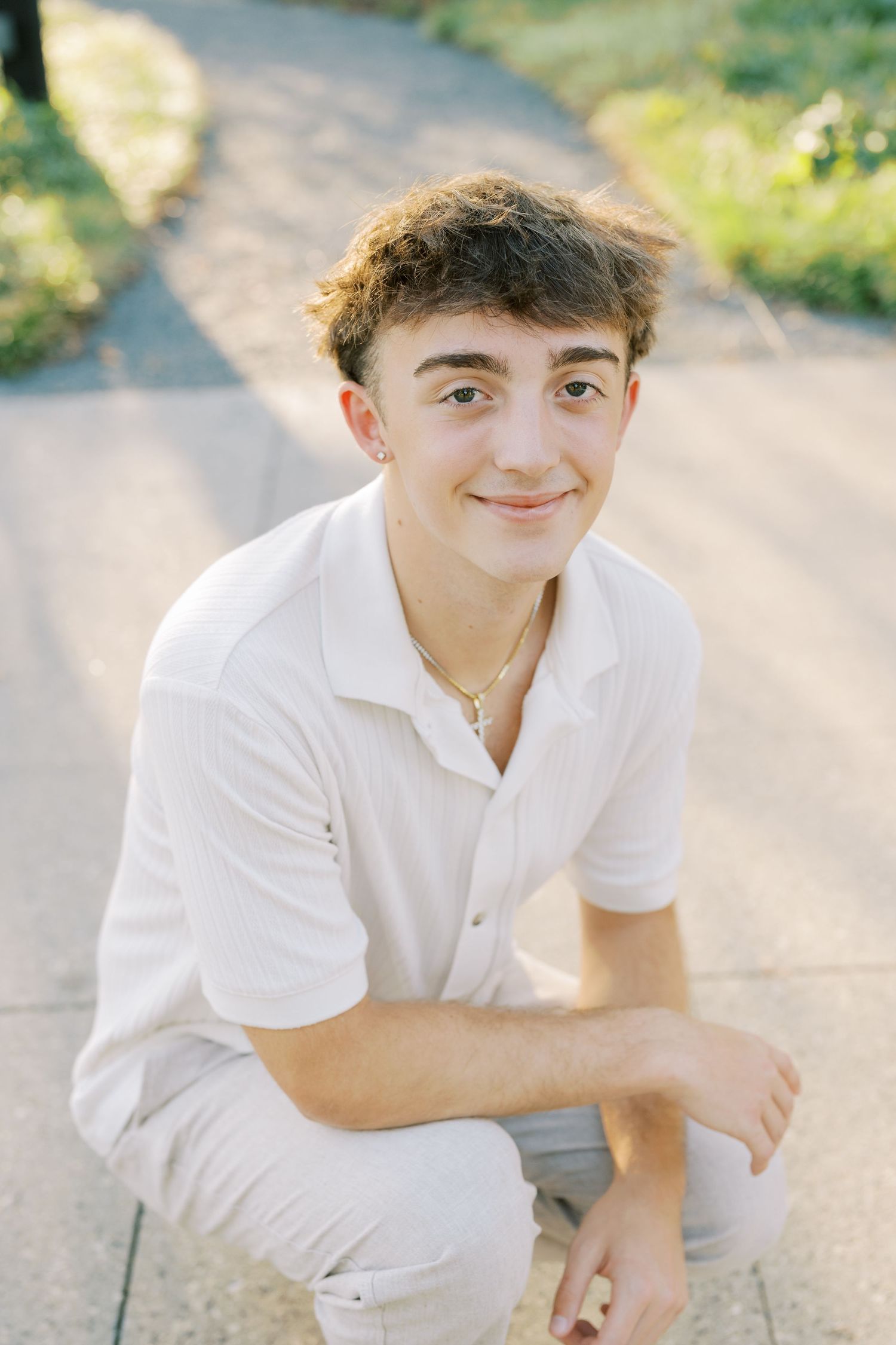 A person in a white polo shirt crouches outdoors in natural lighting against a blurred background.