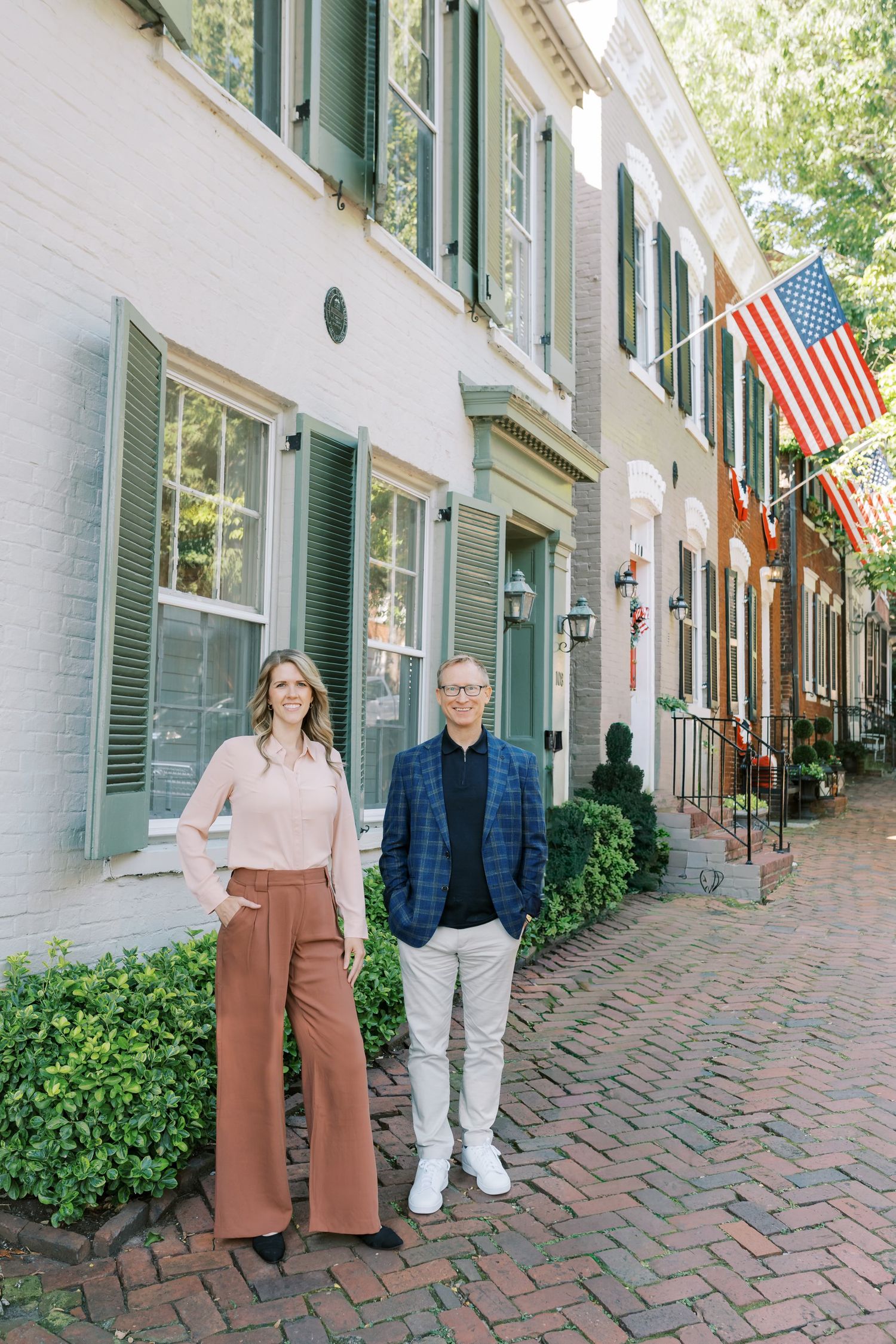 Two people stand on a historic brick sidewalk with American flags and green shuttered buildings in the background.