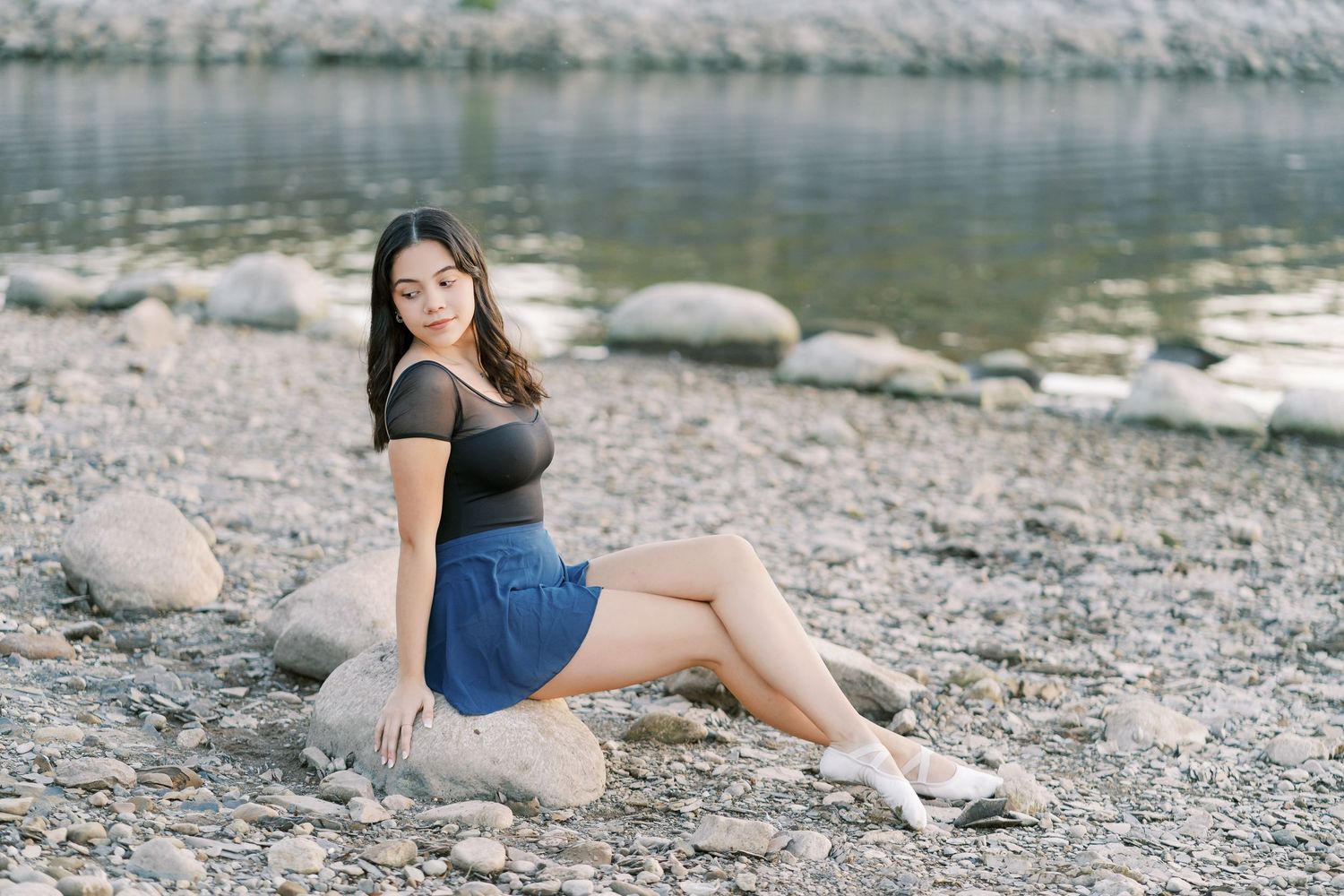 A person in a blue dress poses gracefully on a rocky shoreline near calm water on a bright summer day.