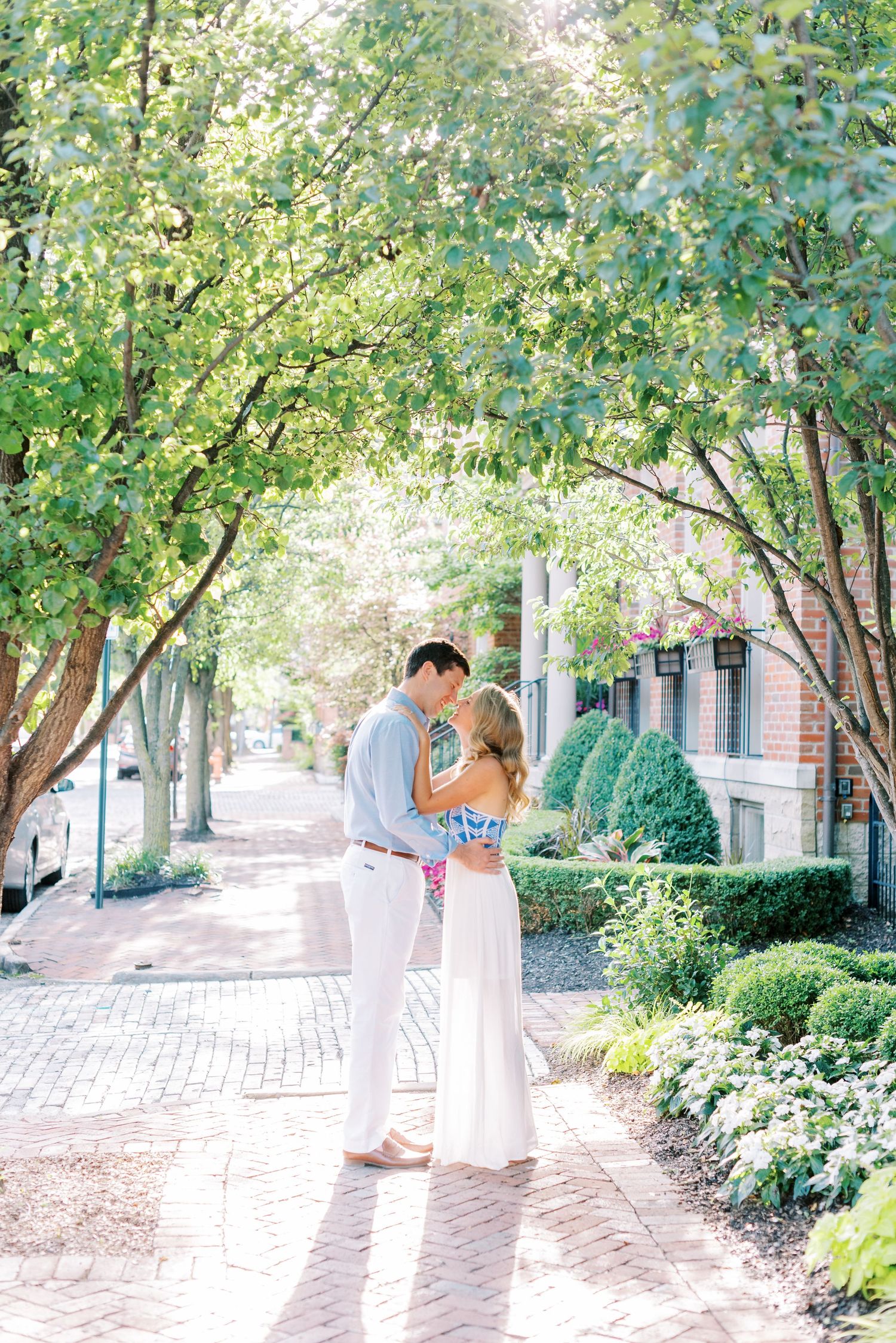 A couple embraces on a sunlit brick pathway surrounded by lush green trees and landscaping in an urban setting.