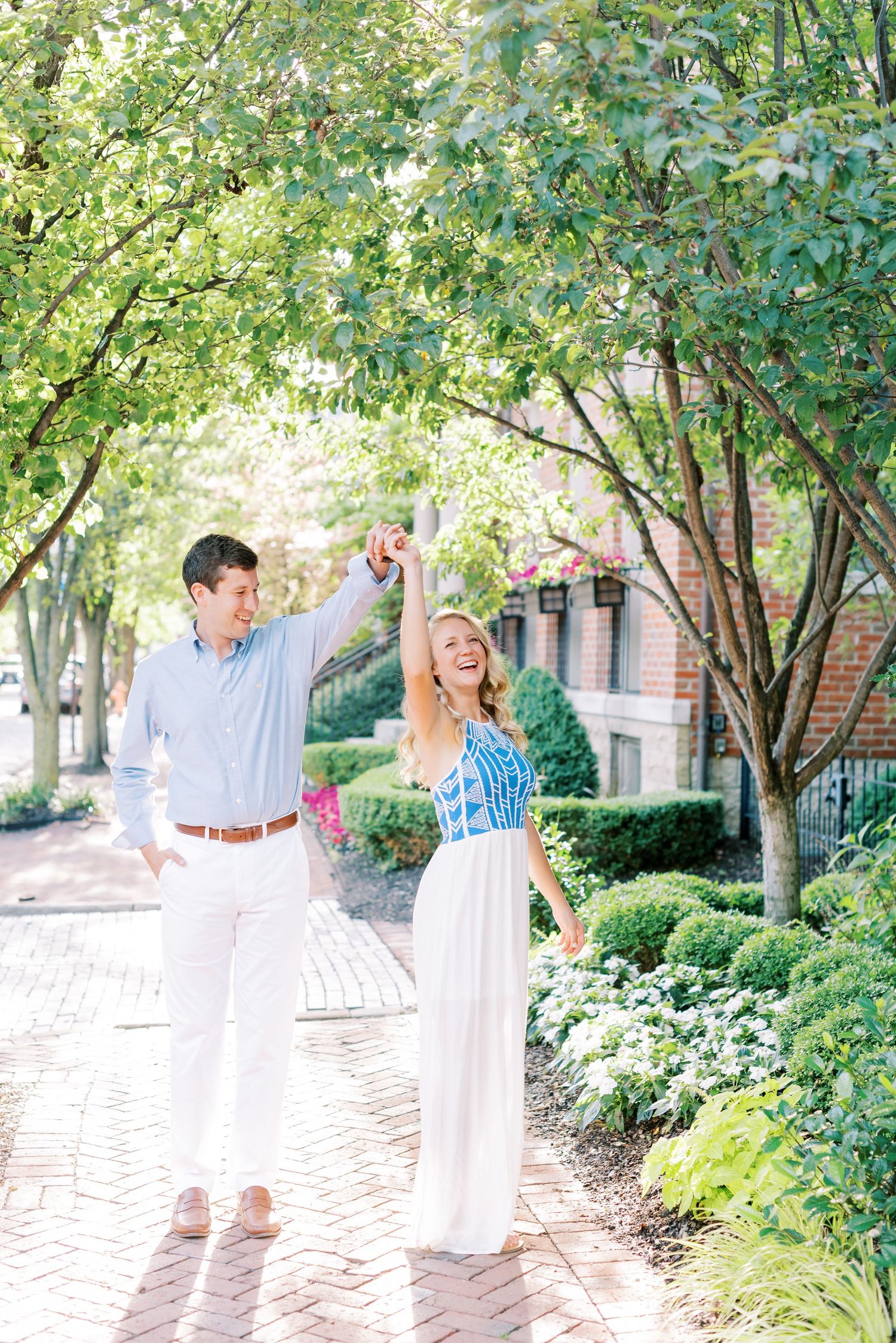 A joyful couple dances together on a brick pathway surrounded by lush green trees and bushes on a sunny day.