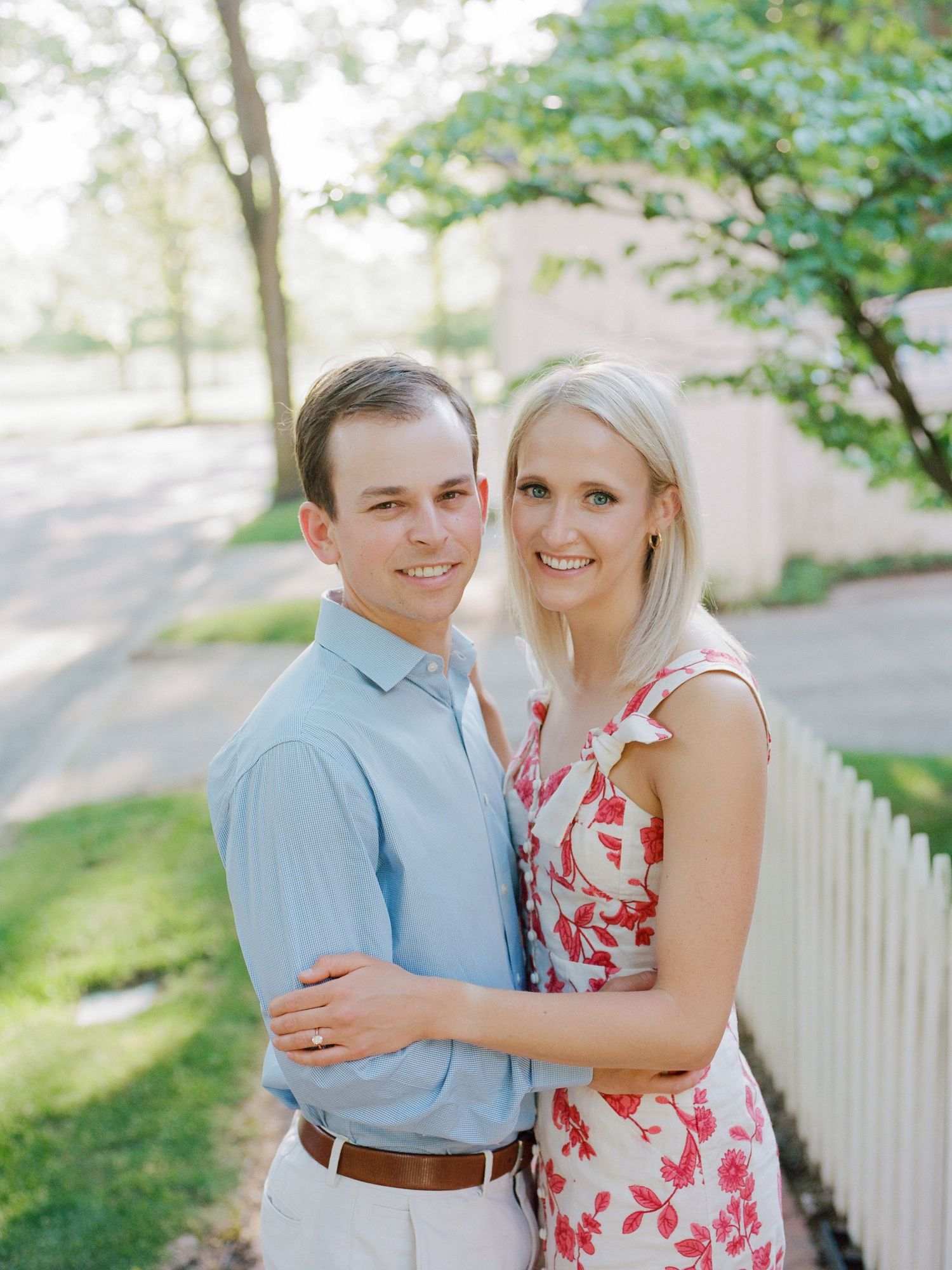 A couple embraces and smiles together during an outdoor engagement photo session near a white picket fence.