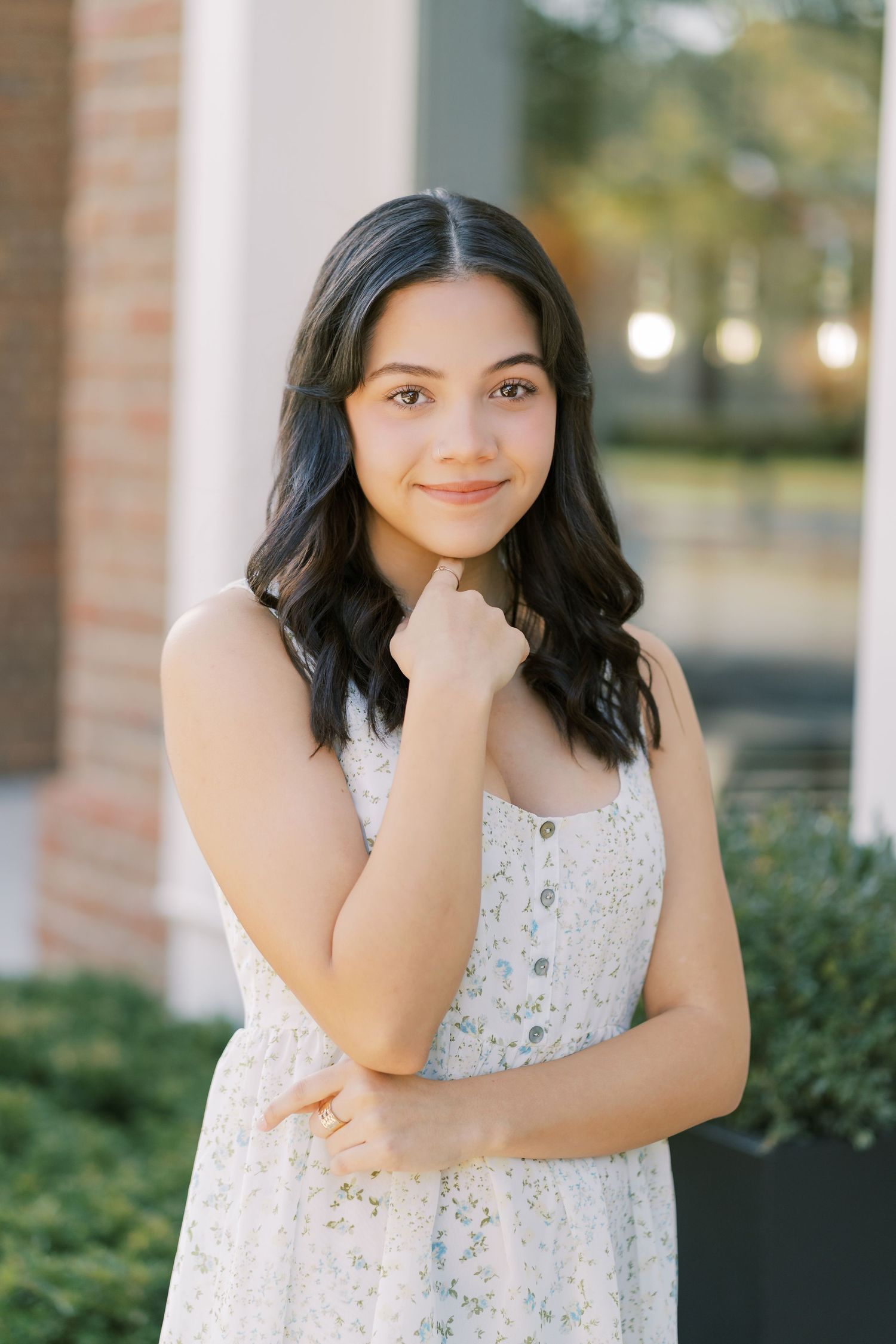 A person in a white lace dress poses outdoors against a backdrop of greenery and brick walls.