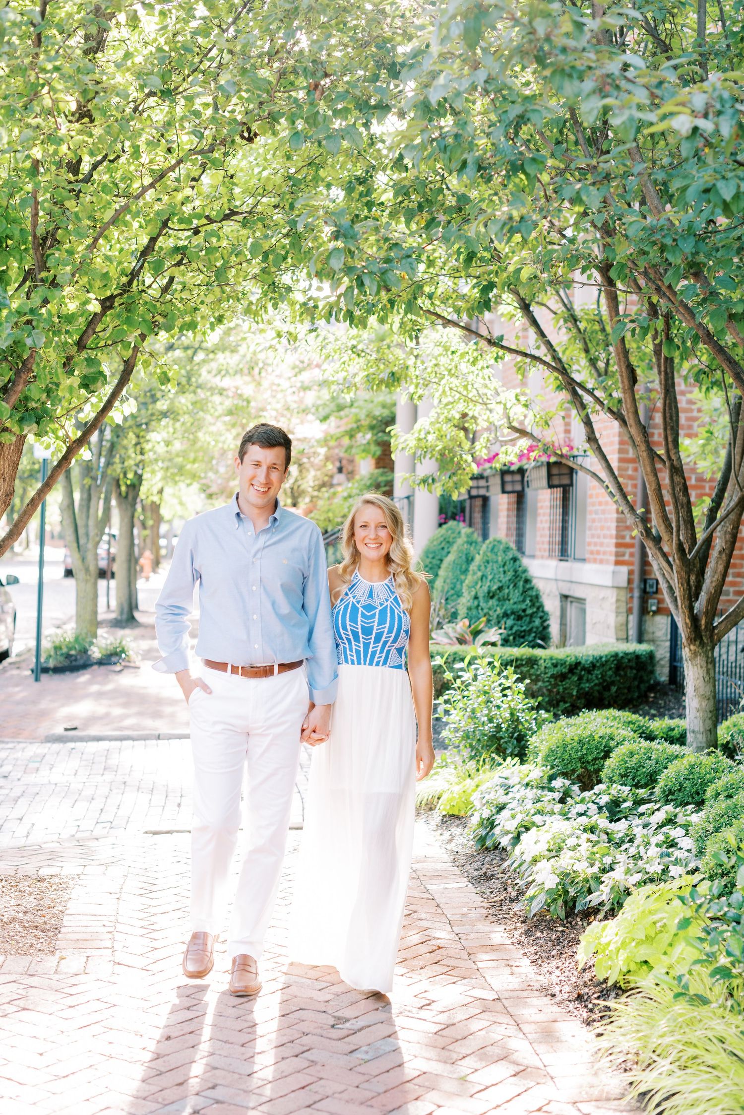 A couple walks together along a brick pathway lined with lush green trees and landscaping on a sunny day.
