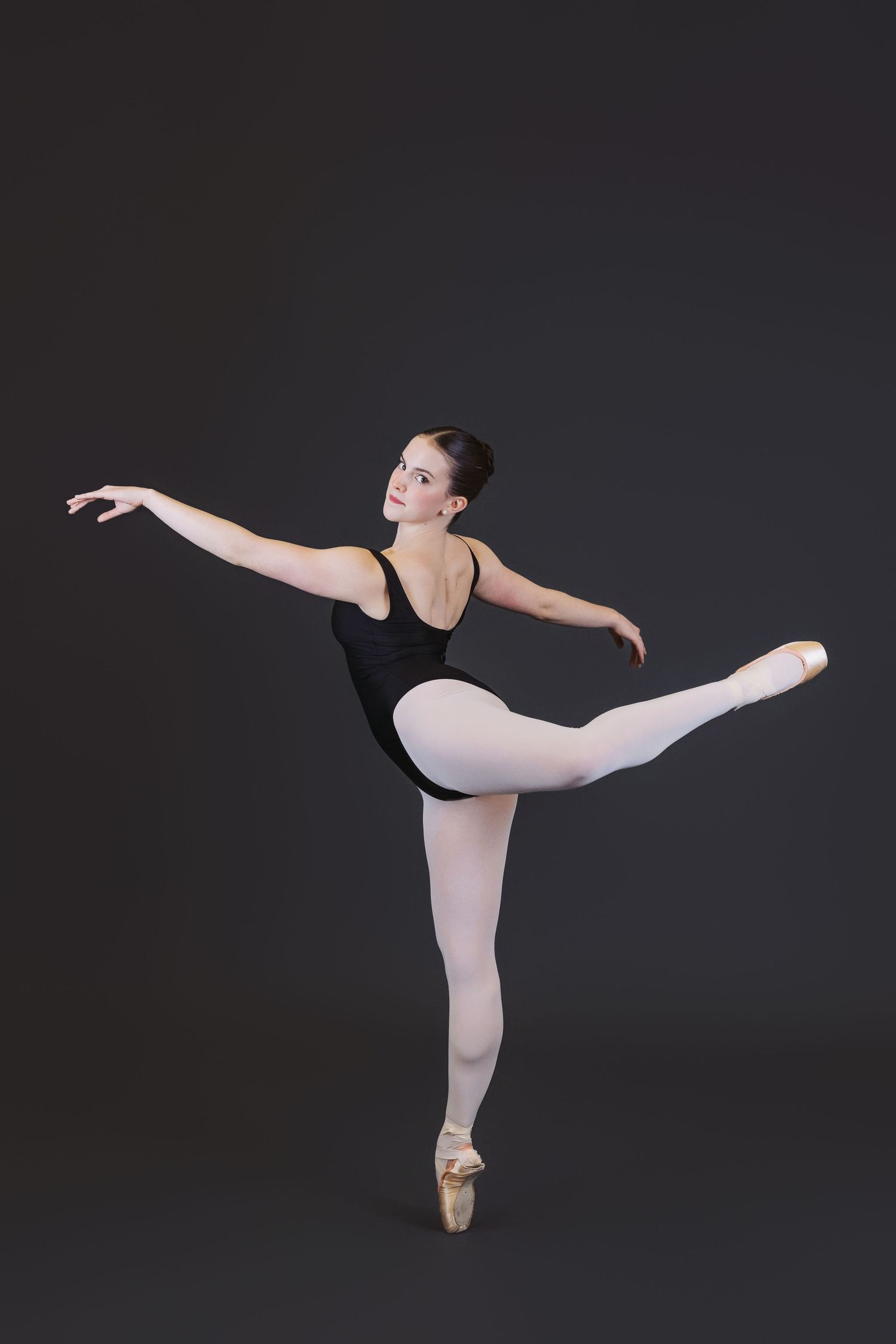 Ballet dancer in black leotard and pink tights performs graceful arabesque pose on pointe against dark background.