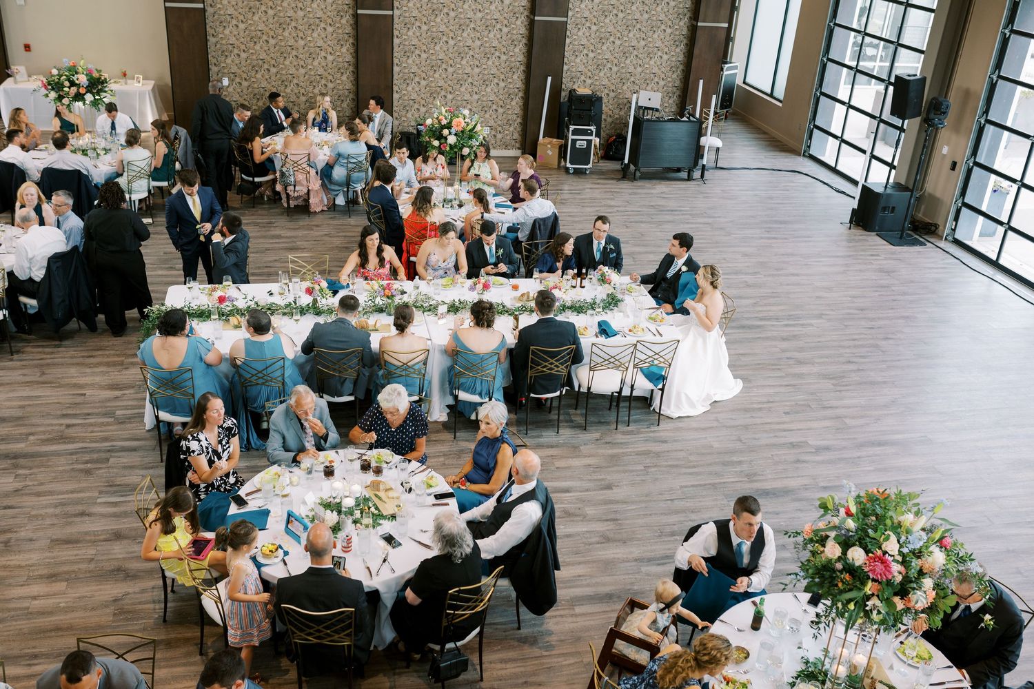 Wedding reception guests seated at round tables in a modern venue with large windows and floral centerpieces.