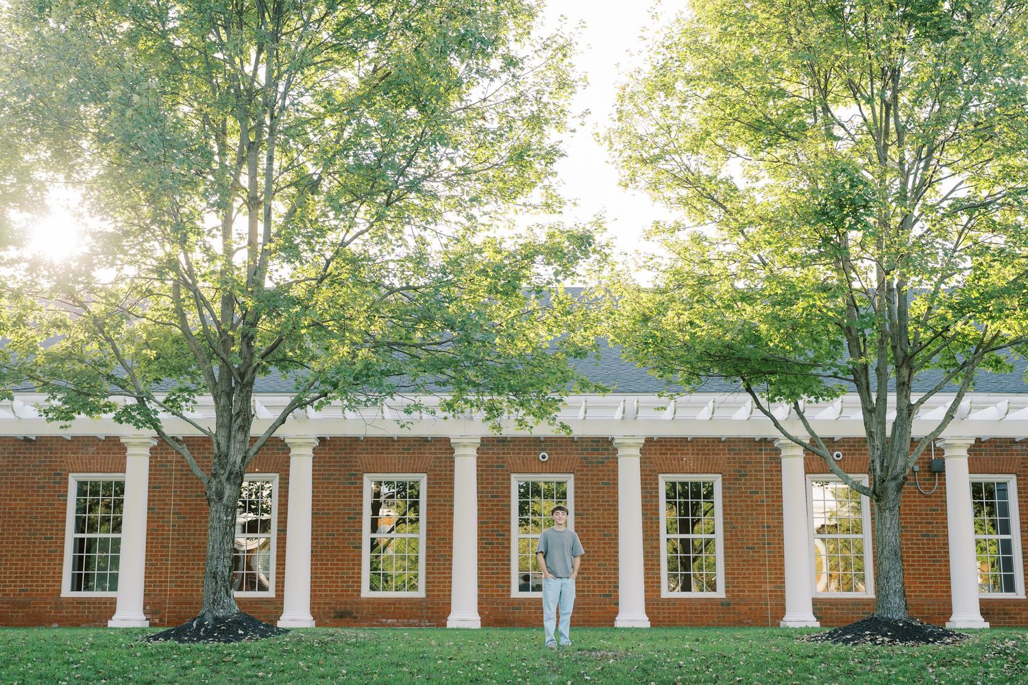 Long brick building with white columns and windows surrounded by green trees in sunlit campus setting.
