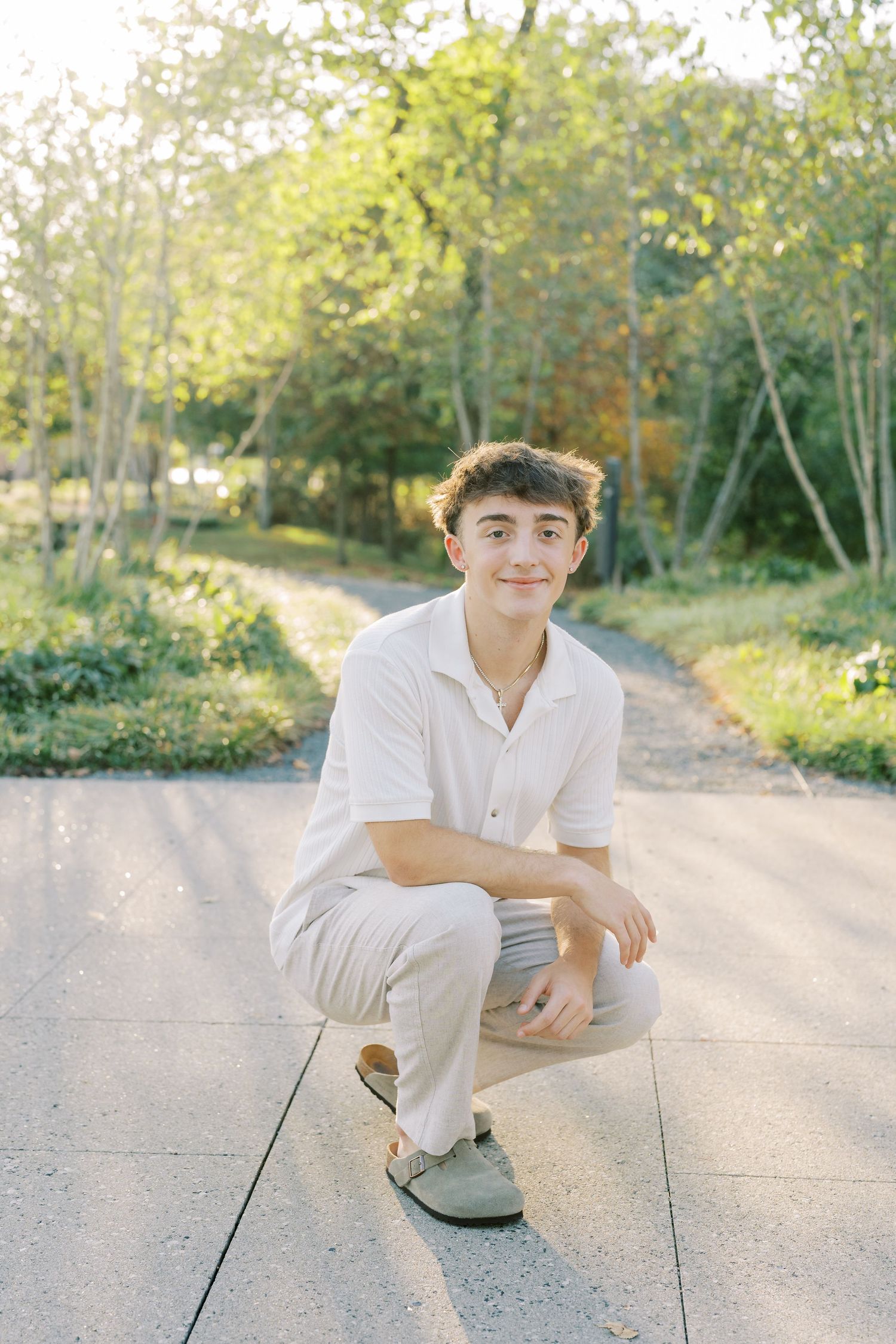 A person wearing white clothing poses on a paved walkway with trees and natural lighting in the background.