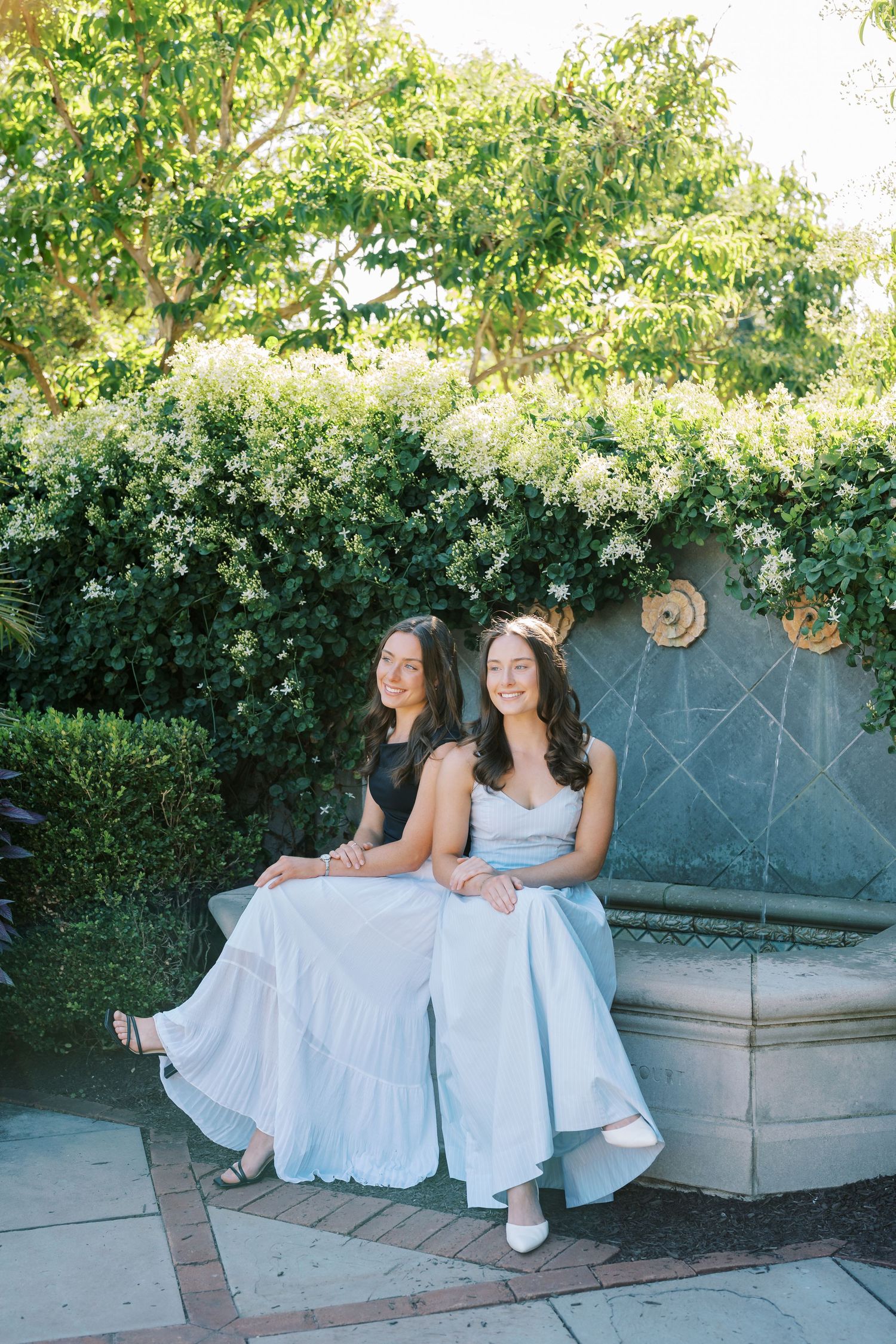 Two people in white dresses sitting together on a stone bench in a garden setting with blooming flowers.