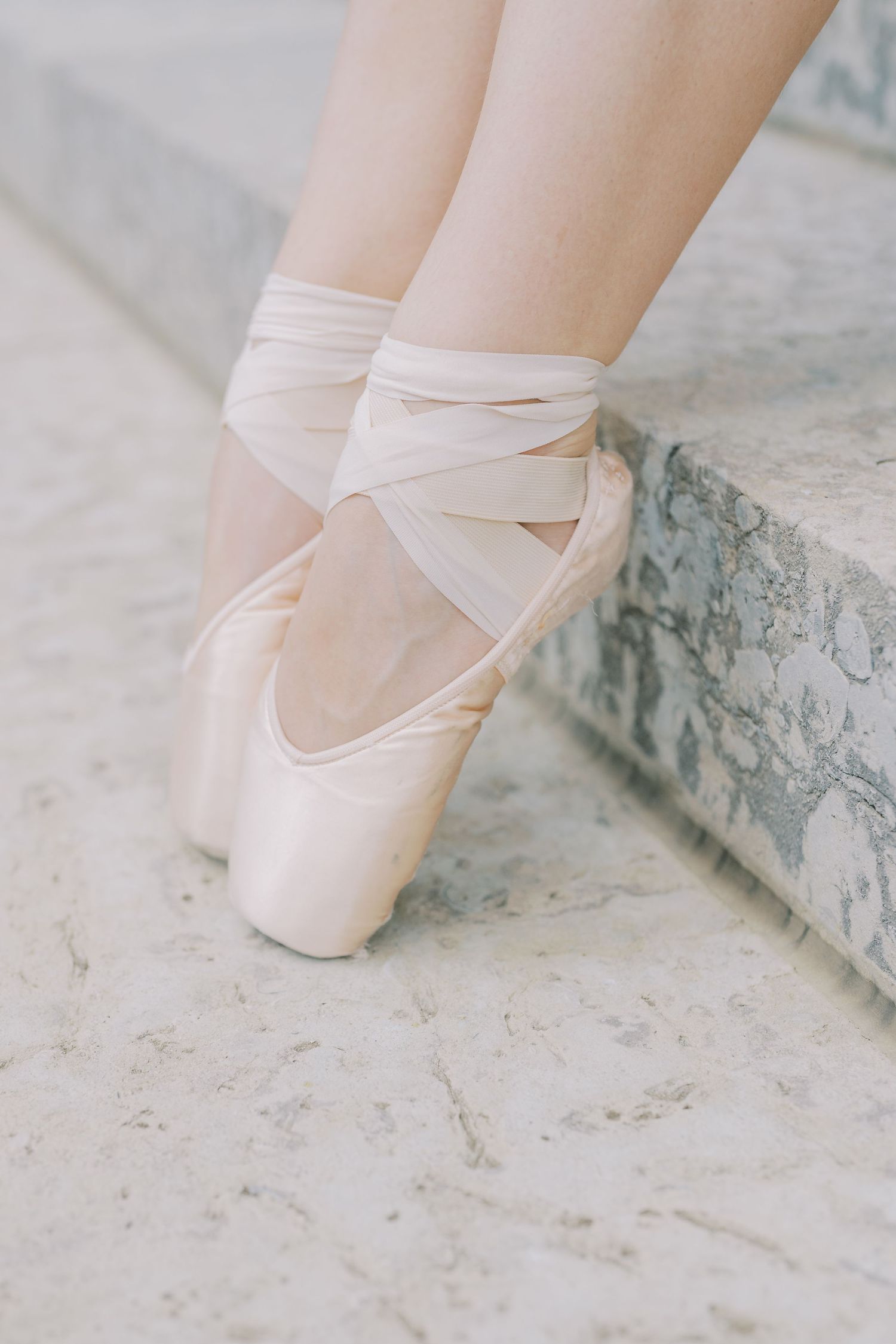 Close-up of pink ballet pointe shoes with satin ribbons tied around ankles on a light stone surface.