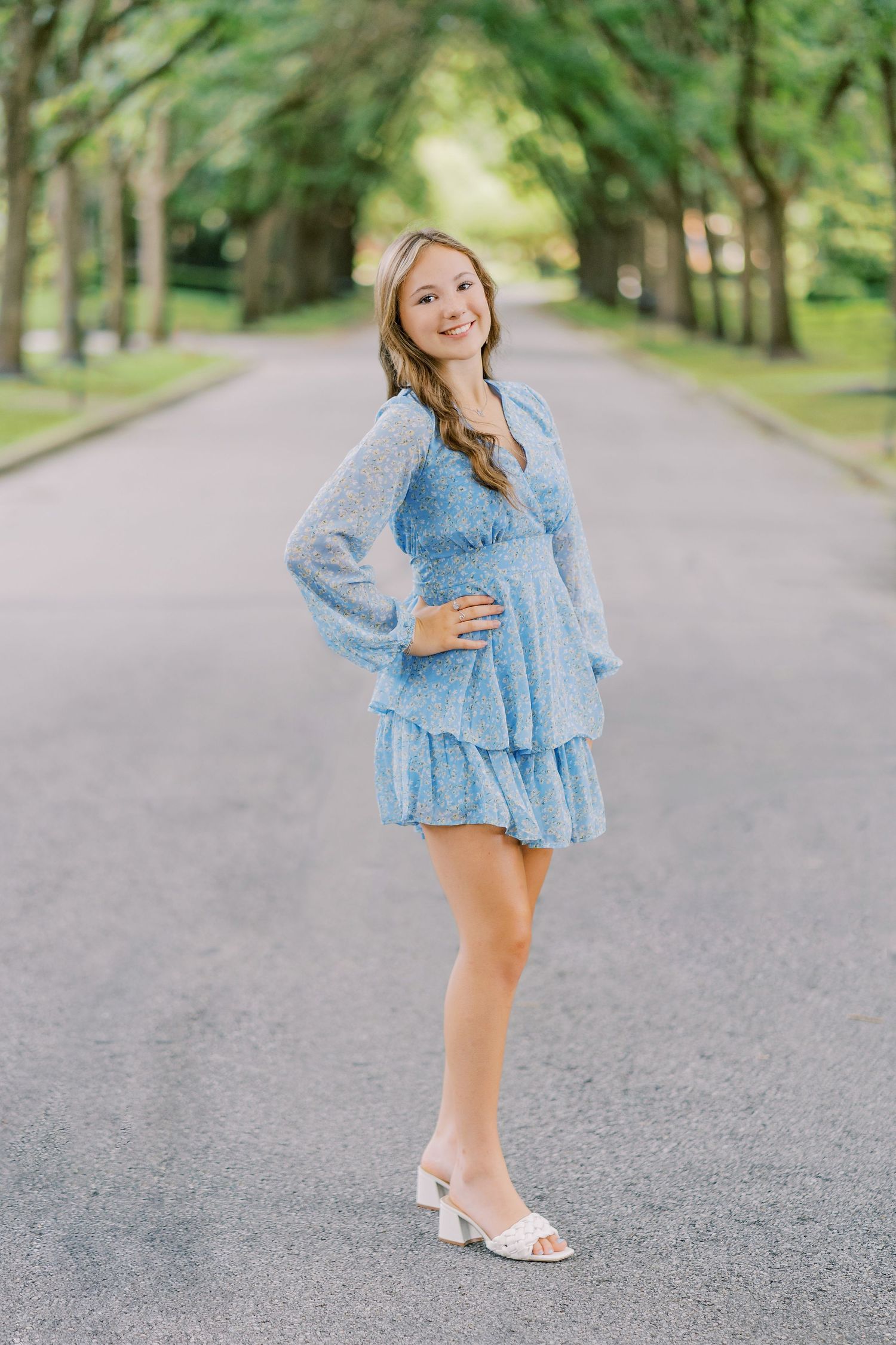 A person in a light blue ruffled dress poses on a tree-lined path wearing white sandals.