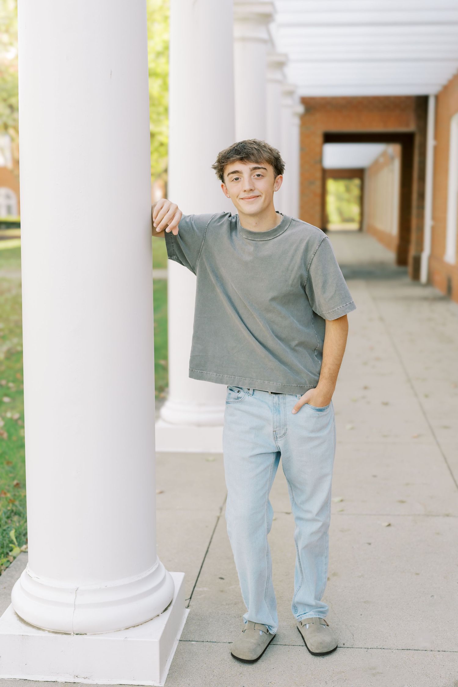 A child in a grey t-shirt and light jeans stands casually leaning against a white column in an outdoor walkway.