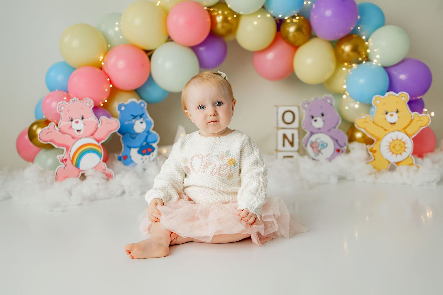 Baby girl in pink tutu sits before colorful Care Bears balloon arch with ONE blocks for first birthday cake smash.