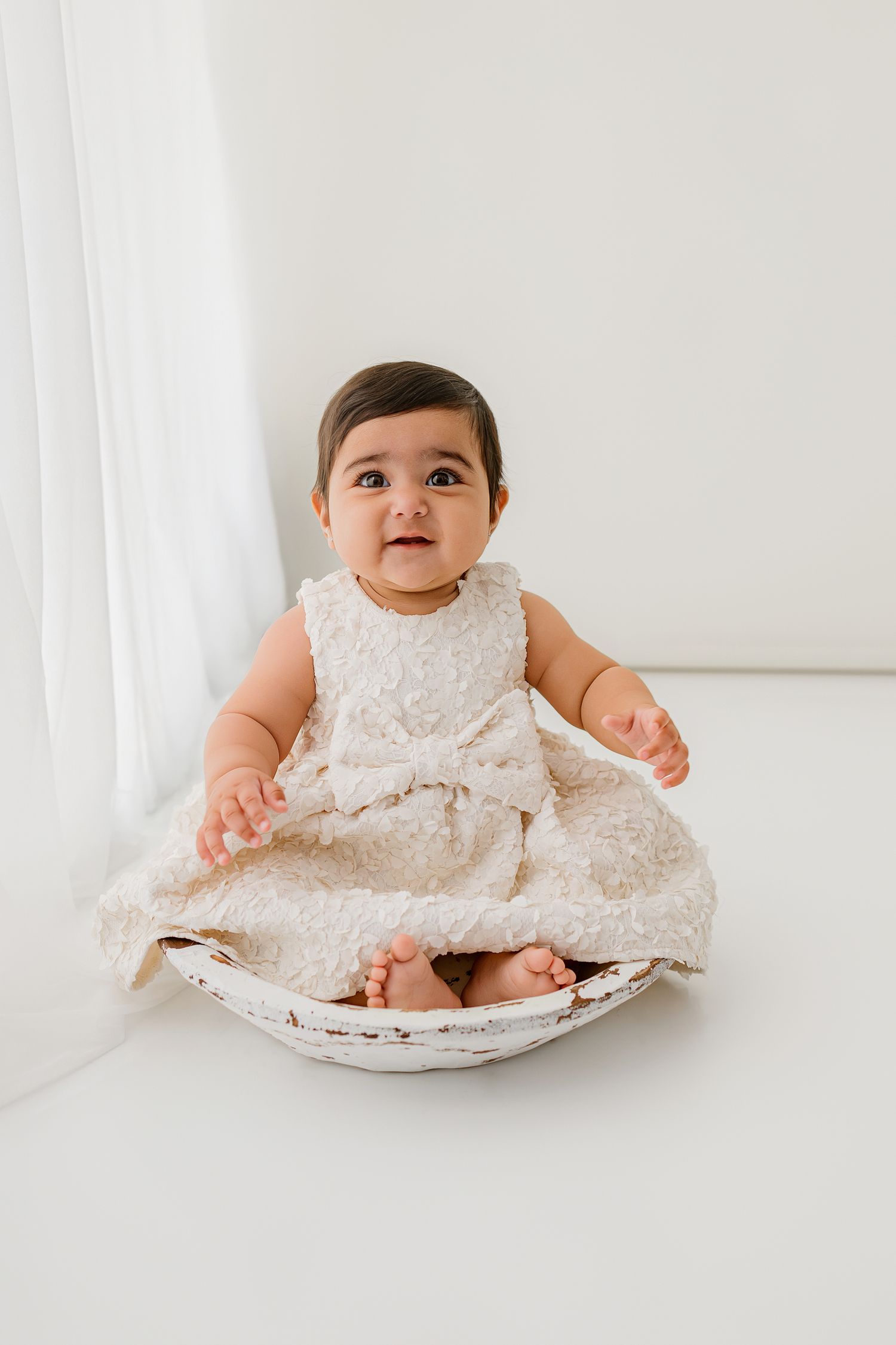 A cute baby girl in a cream lace dress sits in a white bowl prop during a professional studio portrait session.