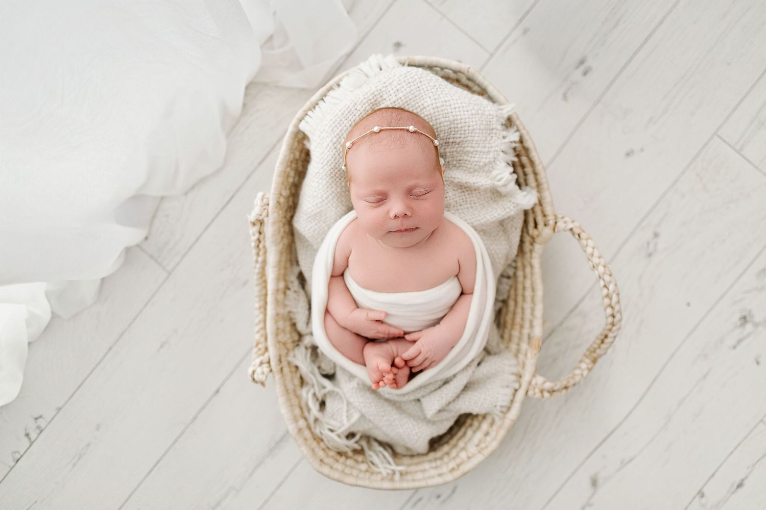 Newborn baby sleeping peacefully in a wicker Moses basket, wrapped in soft white blankets with a delicate headband.