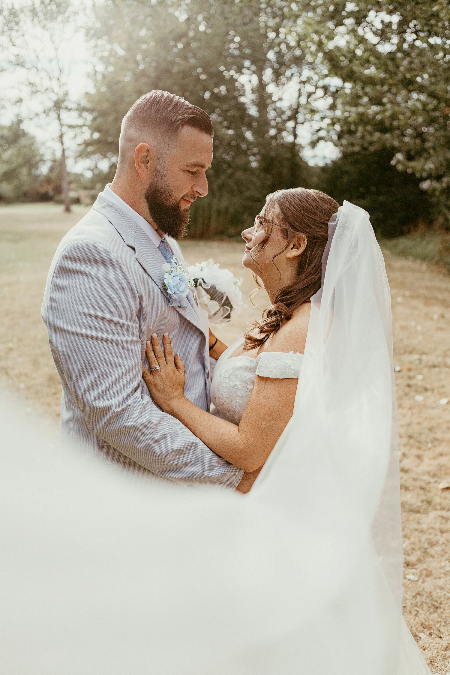 Bride and groom embrace outdoors in golden sunlight, her veil flowing, both smiling joyfully in a lush green garden setting.