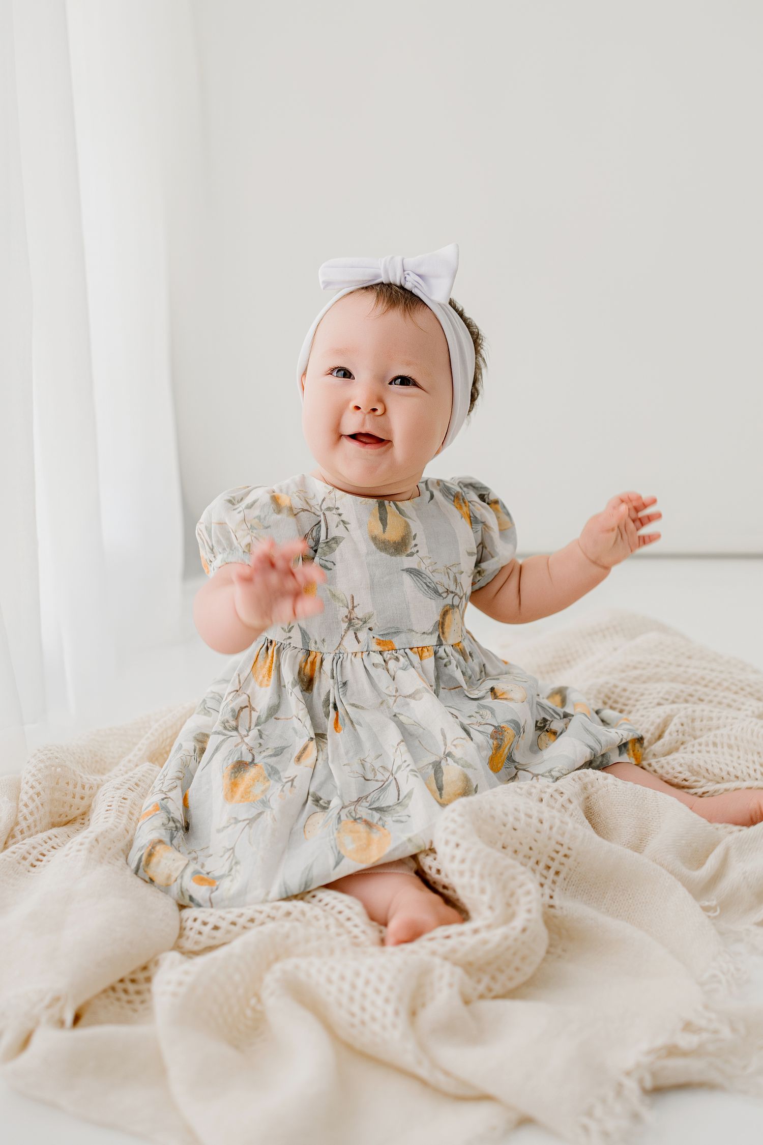 Smiling baby girl in floral dress and white bow headband sitting on cream knit blanket.