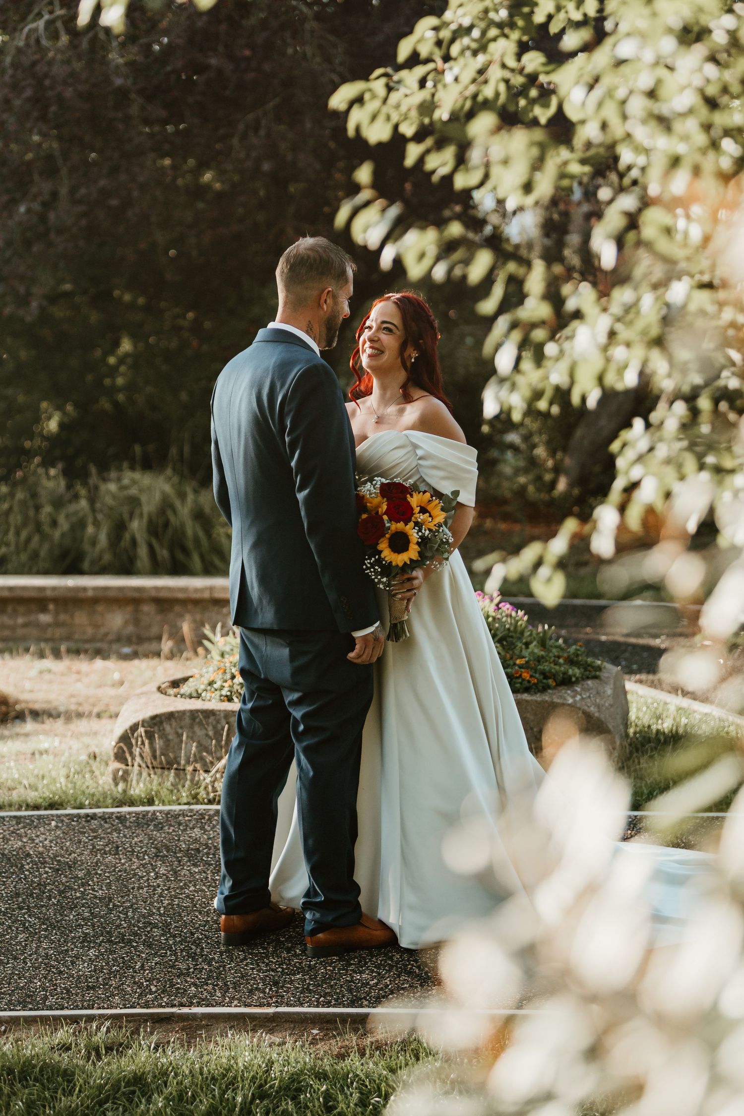 Bride and groom share a joyful moment outdoors, bride holding vibrant sunflower bouquet in elegant off-shoulder white gown.