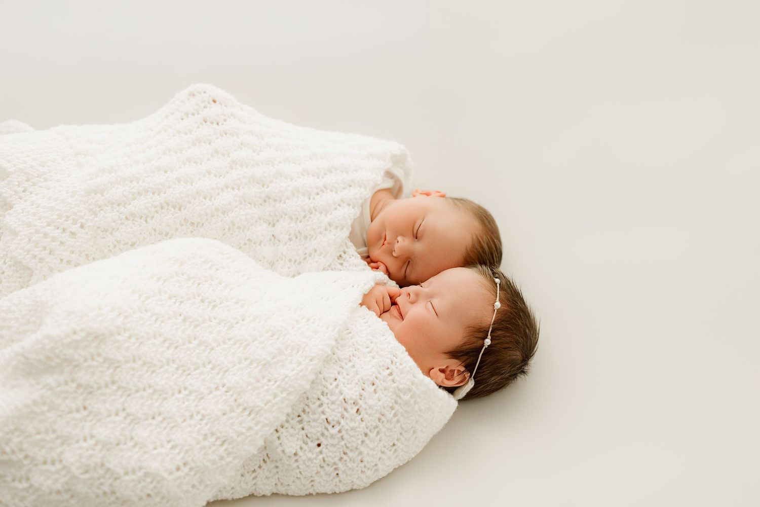 Two sleeping newborn twins swaddled in white knit blankets lying side by side on a white background.