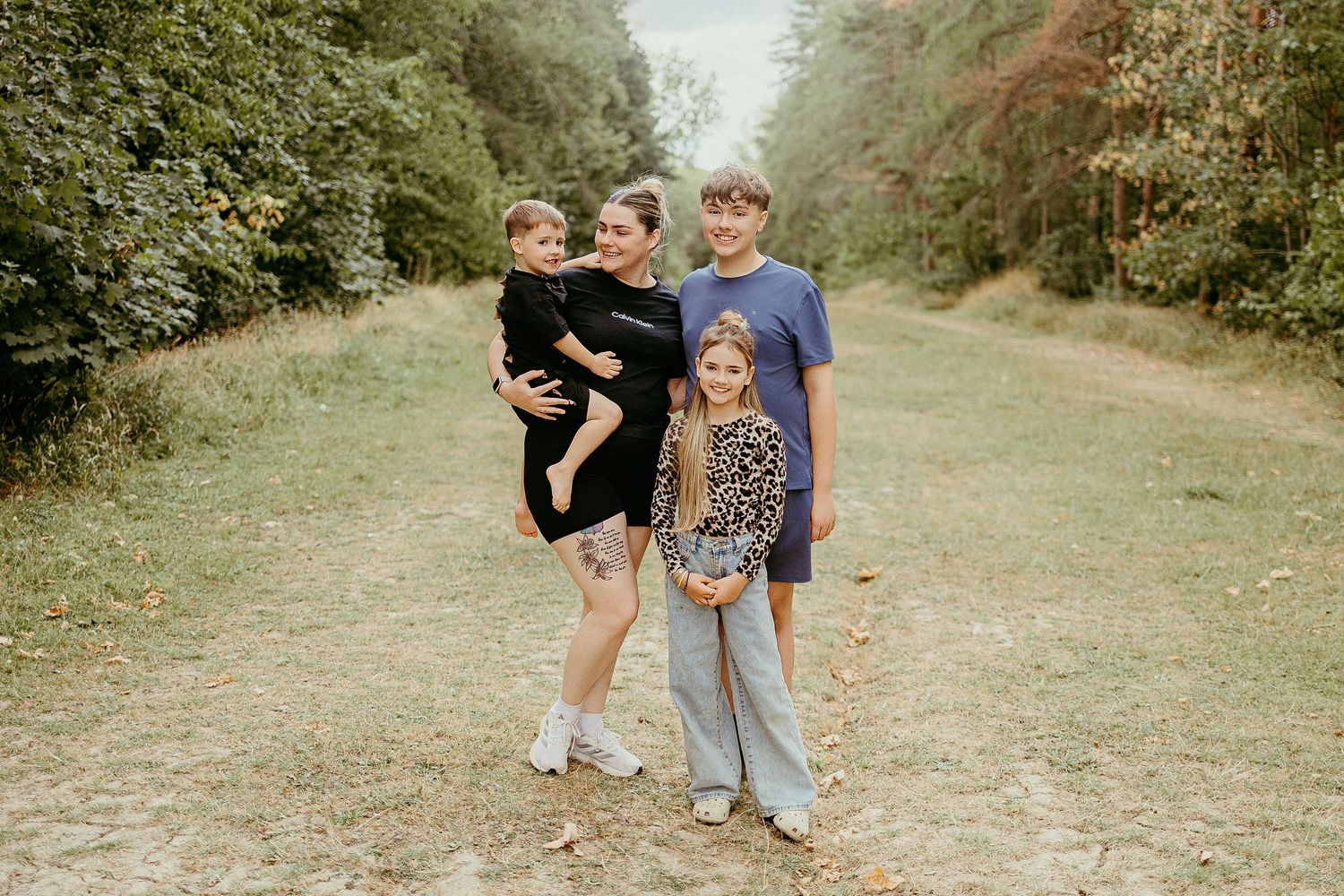Family of four posing on a nature trail surrounded by lush green trees during a fall outdoor photo session.