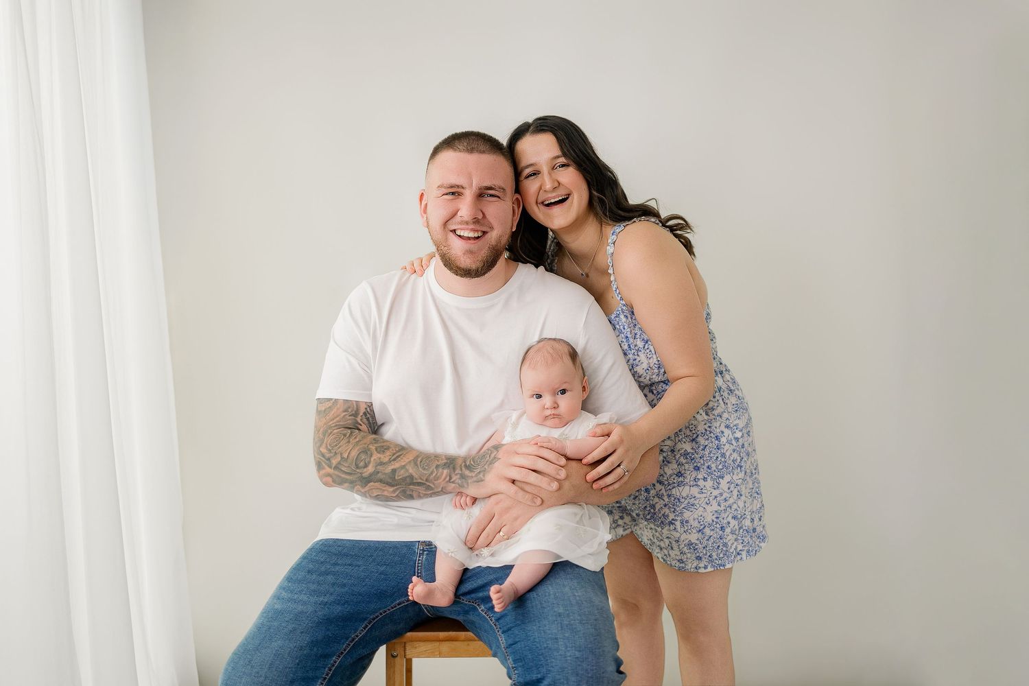 A happy young family with tattoos poses for a studio portrait, parents smiling while holding their newborn baby.