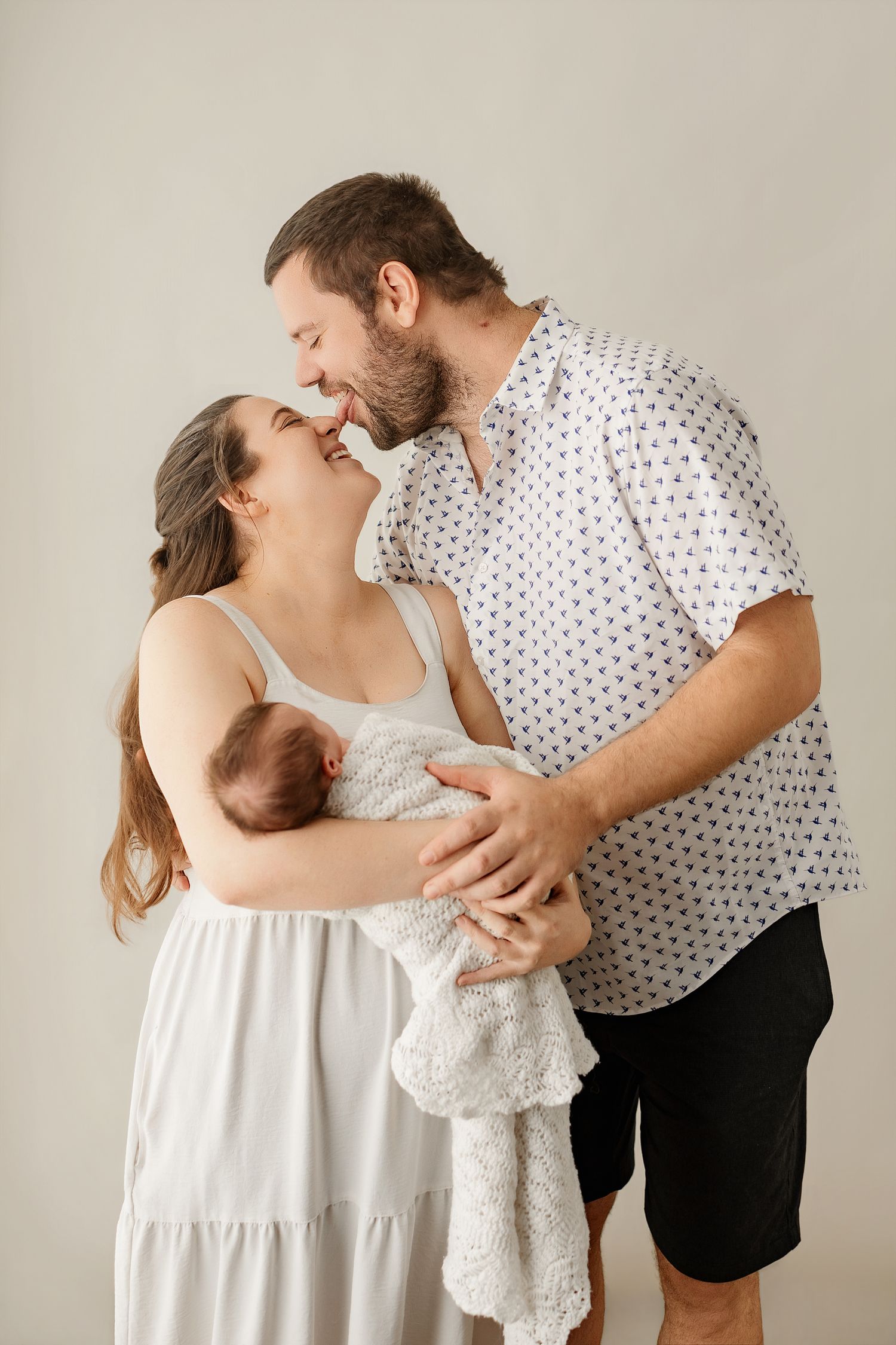 A couple shares a tender kiss while cradling their newborn baby, dressed in white, against a soft neutral background.