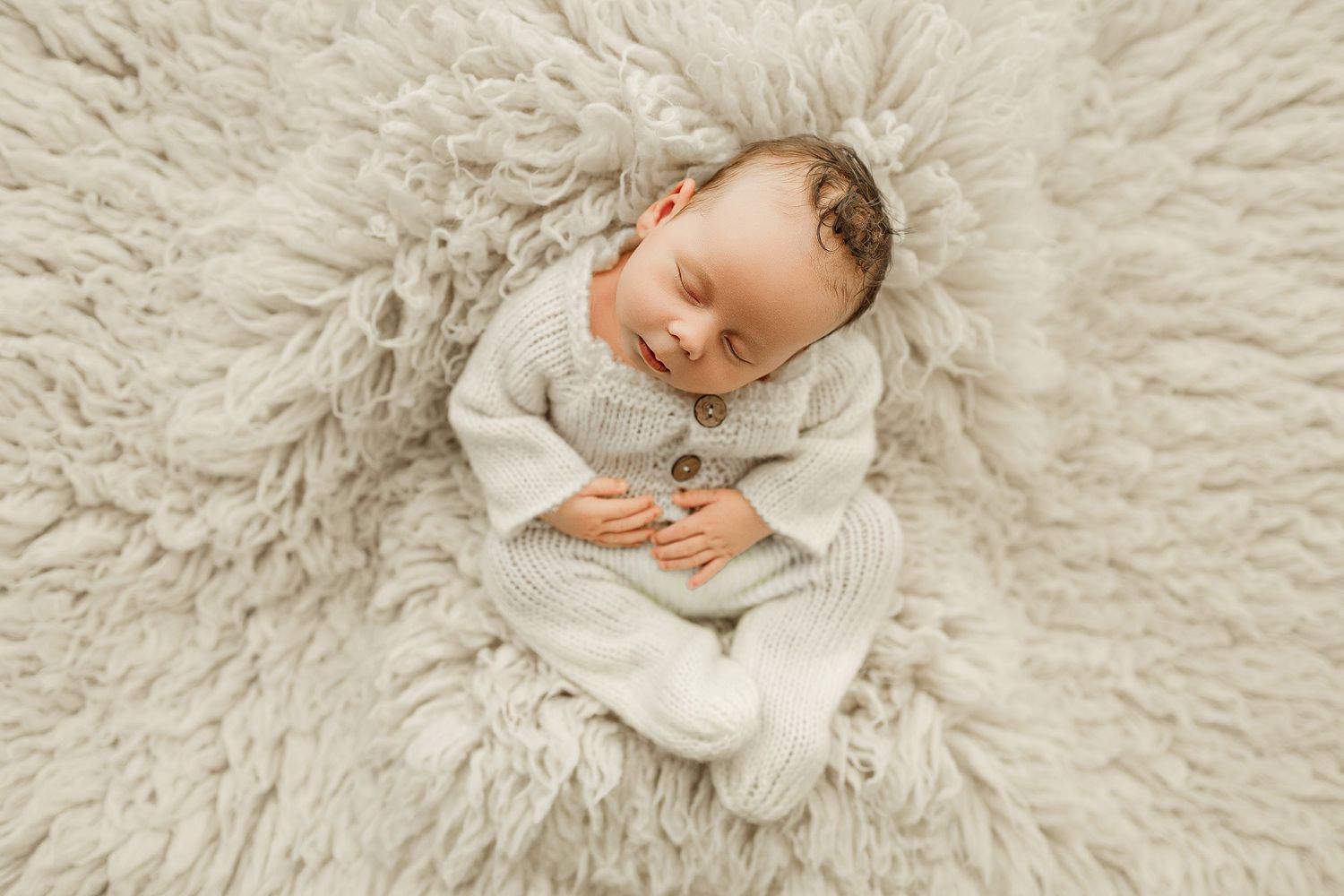 Sleeping newborn baby in cream knit romper curled on fluffy beige rug, peaceful newborn photography.
