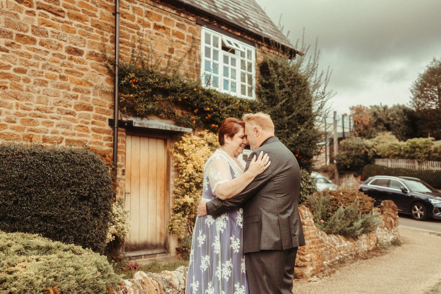 A couple embraces romantically outside a rustic stone cottage with ivy, the woman wearing a floral blue dress.