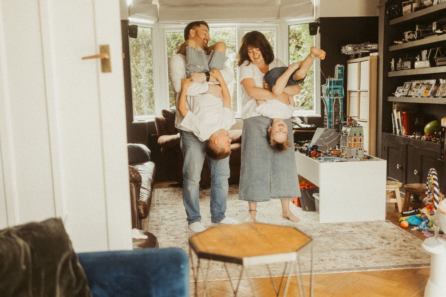 Two parents playfully hold their children upside down in a bright, cozy living room during a fun family moment.