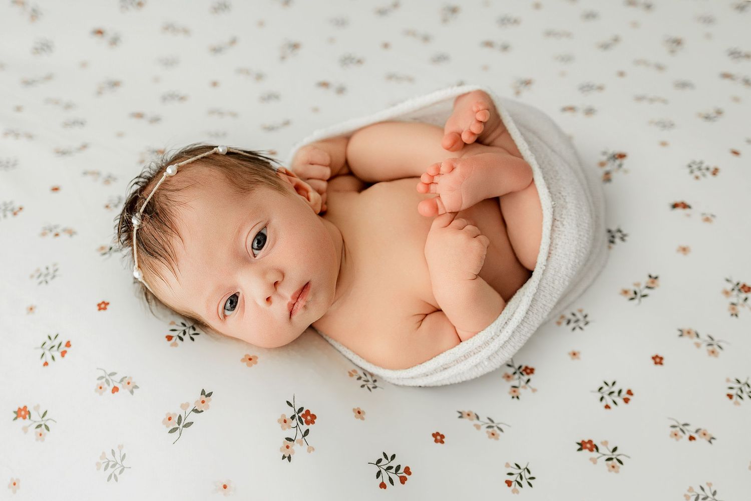 Newborn baby wrapped in white swaddle lying on floral fabric, gazing upward during professional newborn photography session.