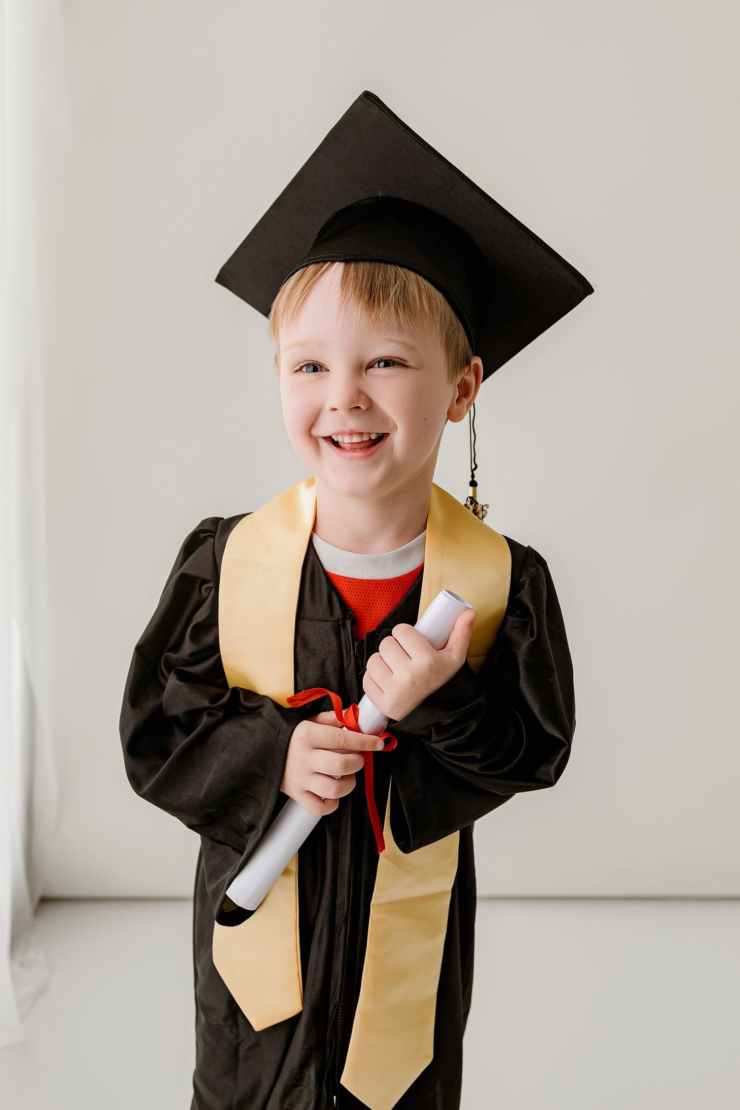 Happy young boy in black graduation gown, cap, and gold stole holding a diploma, smiling at camera.