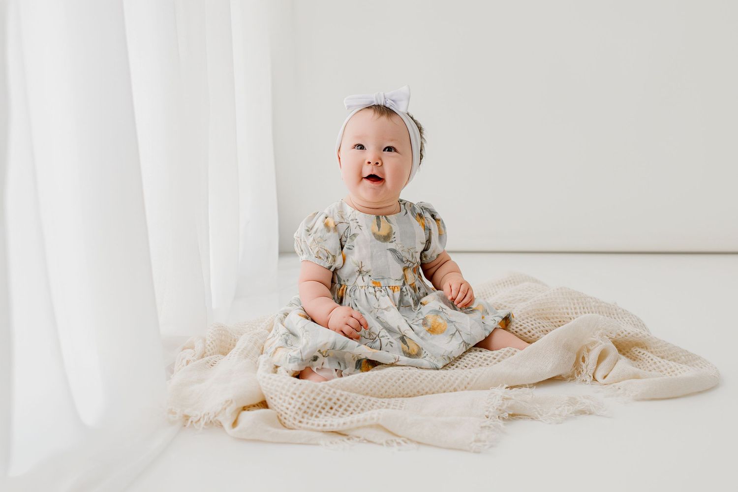 Adorable baby girl in floral dress and headband sitting on cream knit blanket in bright white studio.