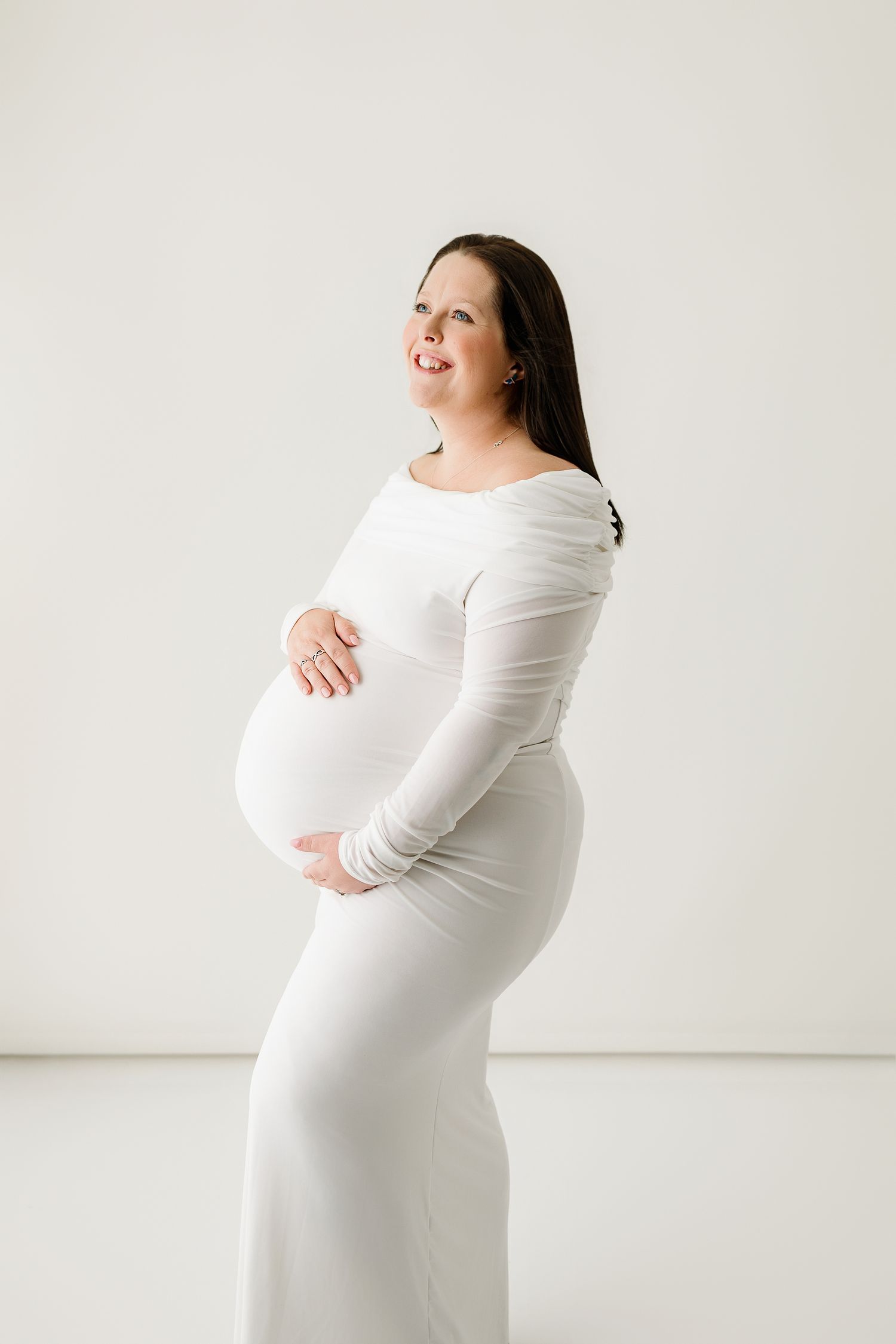 Pregnant woman in white long-sleeve dress cradling her baby bump, smiling against a white background.