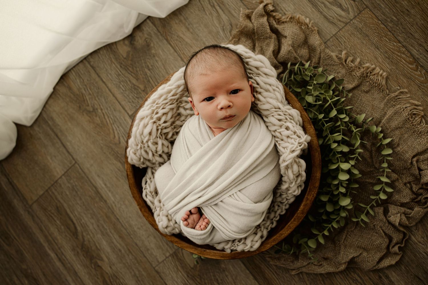 Newborn baby swaddled in white wrap nestled in wooden bowl with chunky knit blanket and eucalyptus leaves.