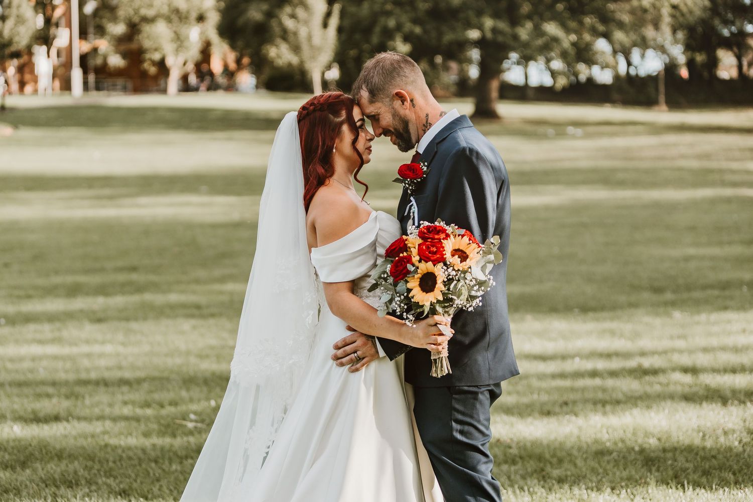 Bride and groom touching foreheads outdoors, bride in white gown with veil, groom in gray suit holding sunflower bouquet.