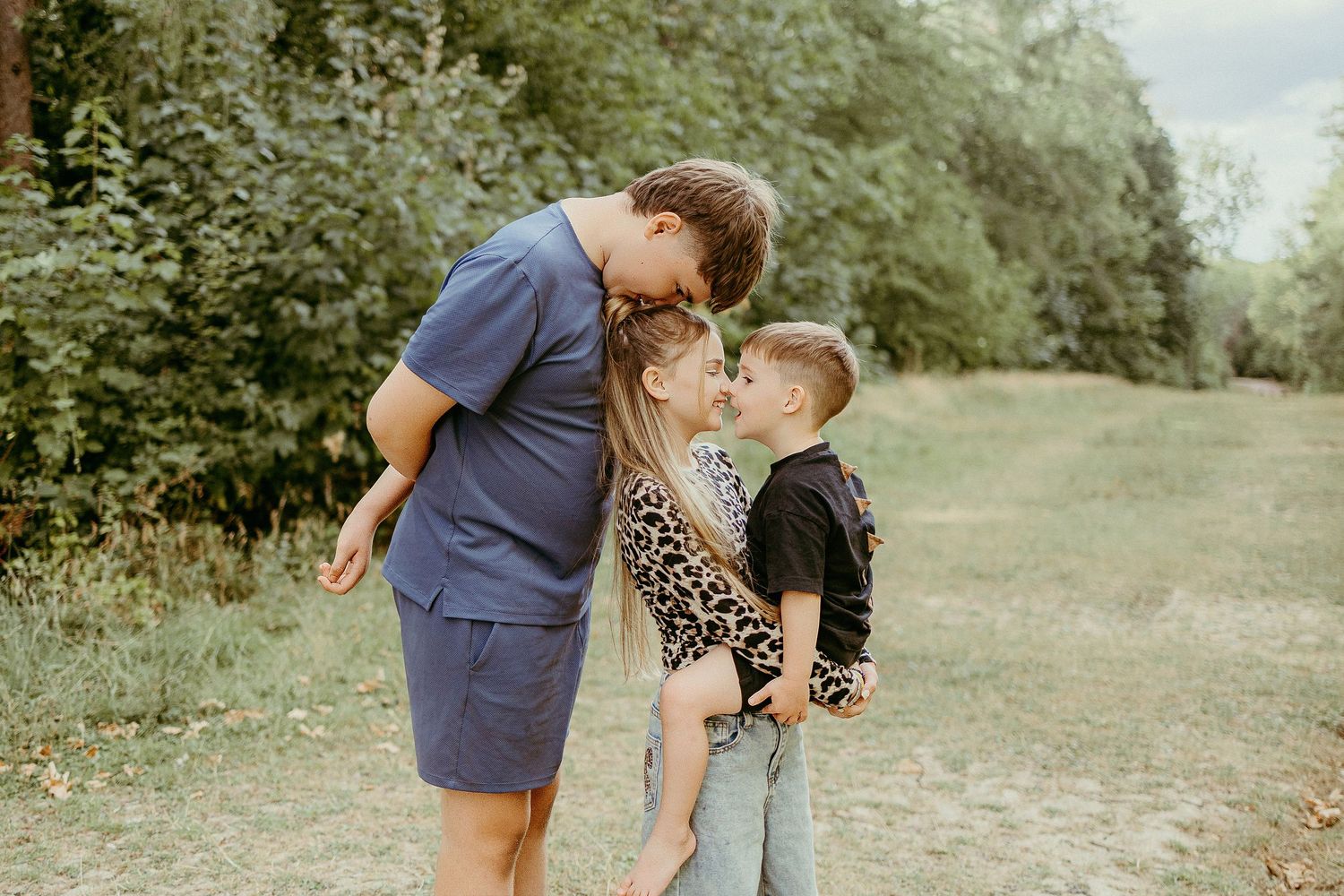 Brother tenderly kissing two younger siblings outdoors in green nature setting.