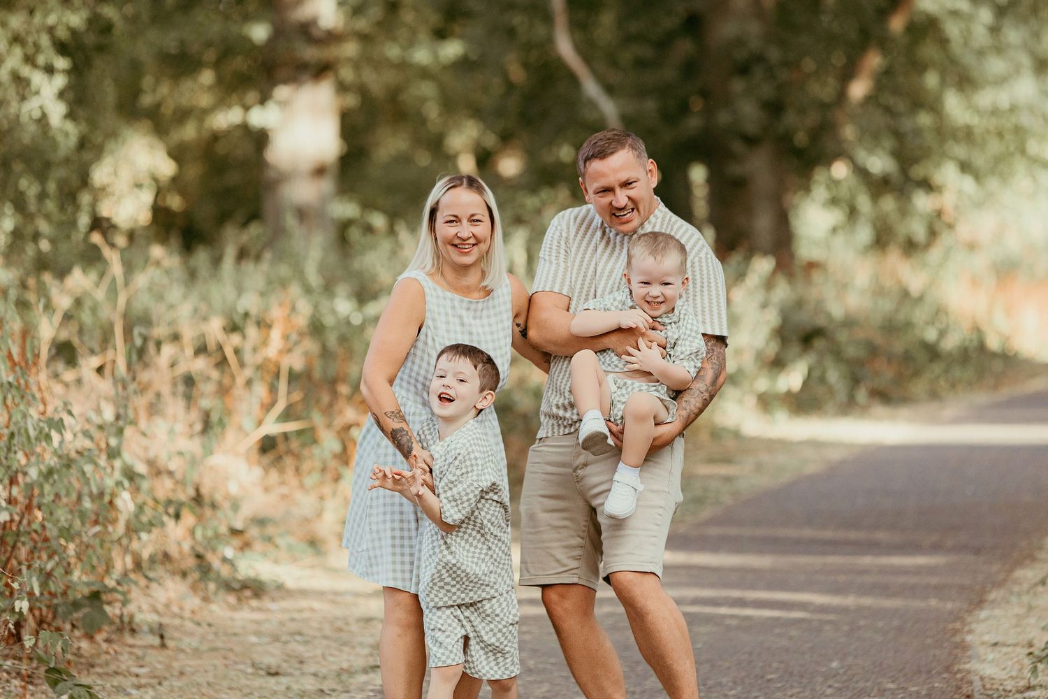 Happy family of four posing outdoors on a path, parents smiling with two young children on a sunny day.