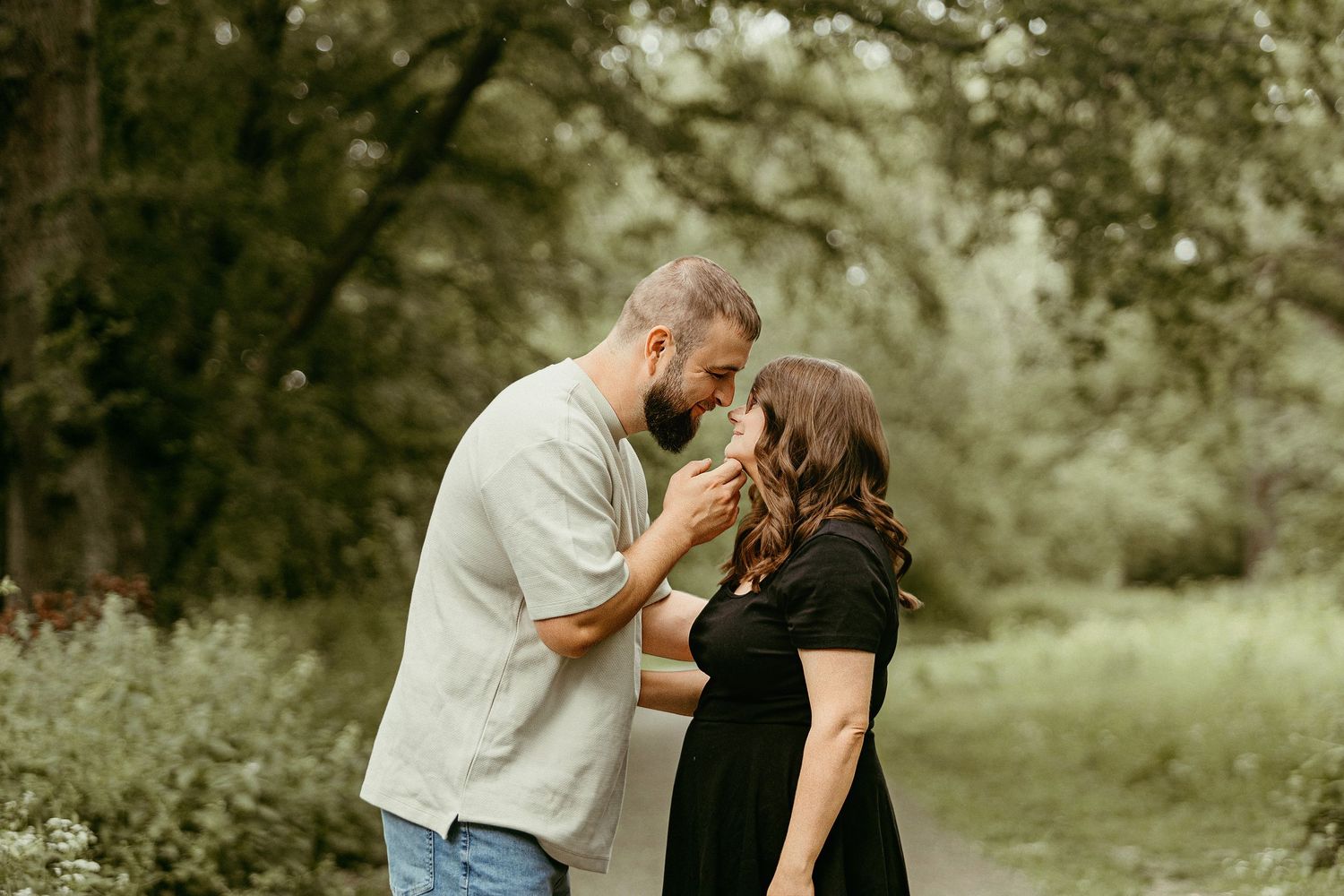 A bearded man tenderly holds a flower to a pregnant woman's nose outdoors in a lush green park setting.