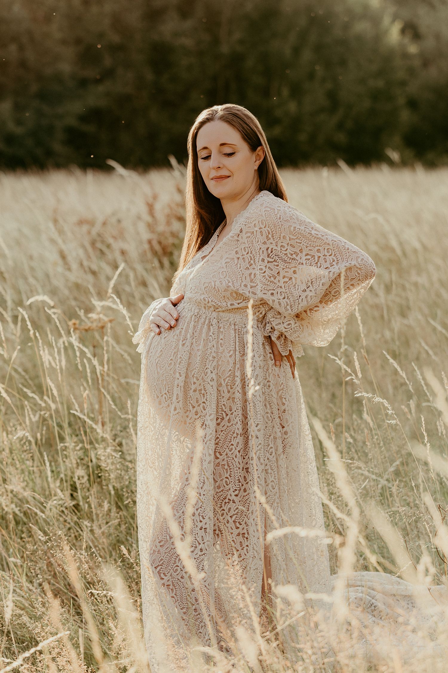 Pregnant woman in lace dress standing in golden field, cradling baby bump during outdoor maternity photo session.