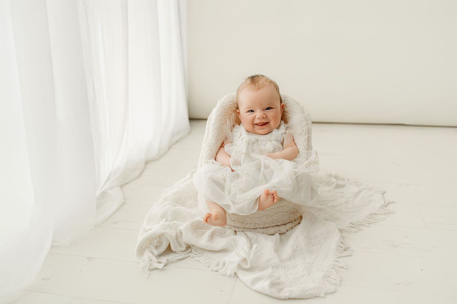 Smiling baby in white romper sitting in a round wooden bowl with white blankets in a bright studio.