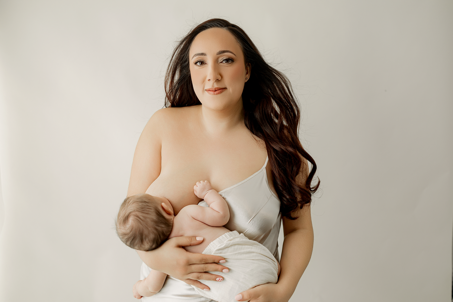 A mother with long dark hair breastfeeding her newborn baby against a soft white background.