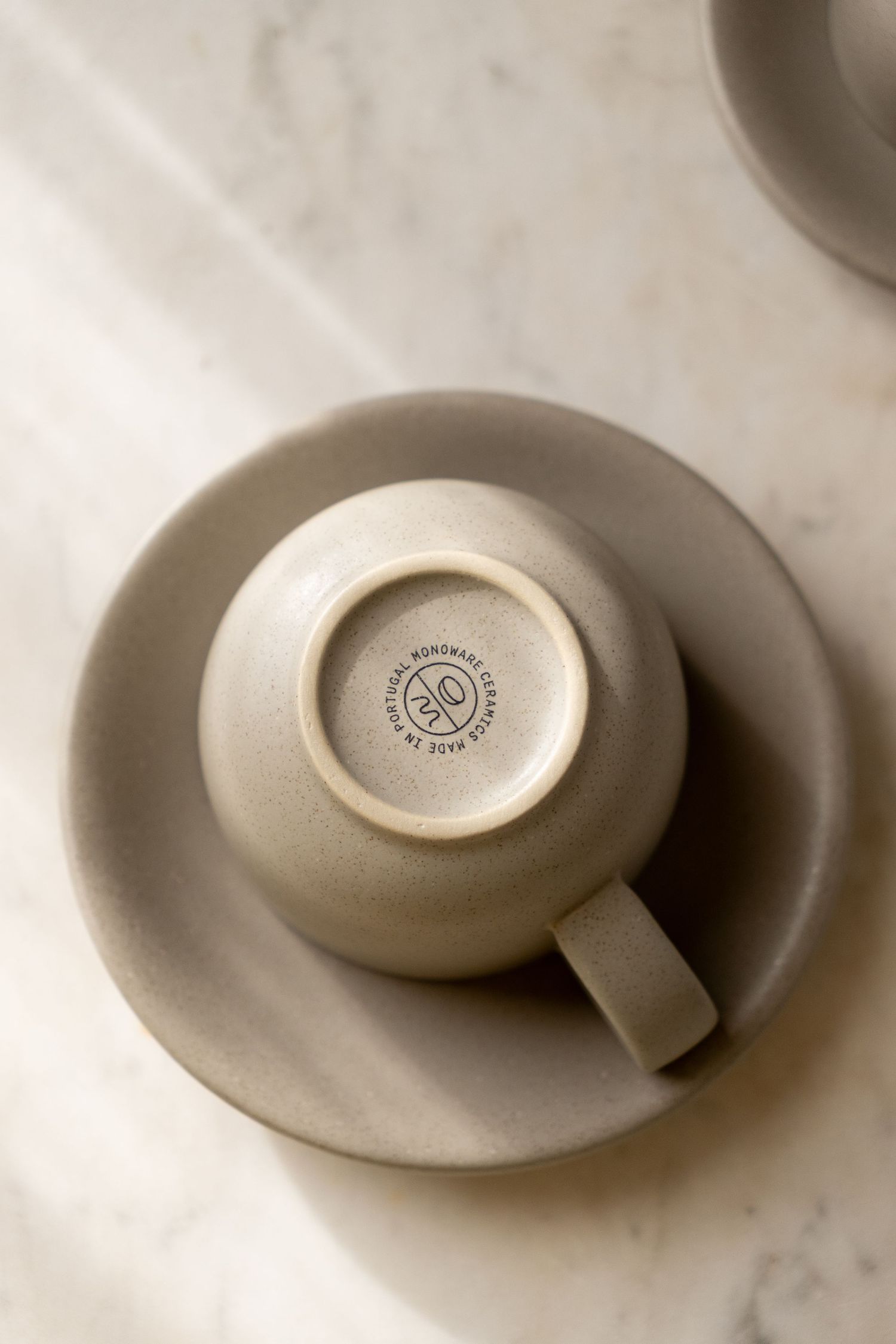 Upturned matte grey ceramic espresso cup on matching saucer, showing maker's mark stamp on base, marble surface.