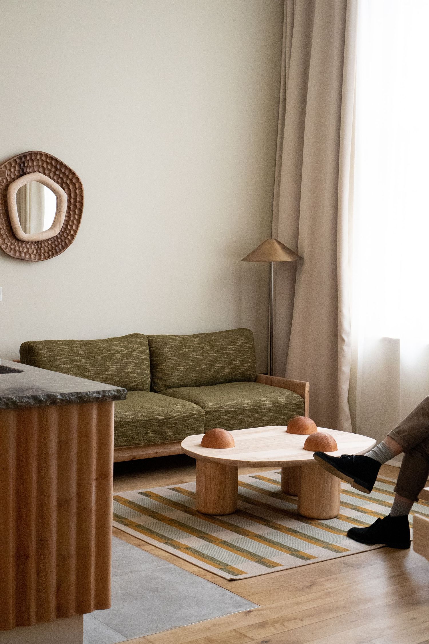Modern living room with green sofa, wooden coffee table, striped rug, rattan mirror, and warm floor lamp by window.
