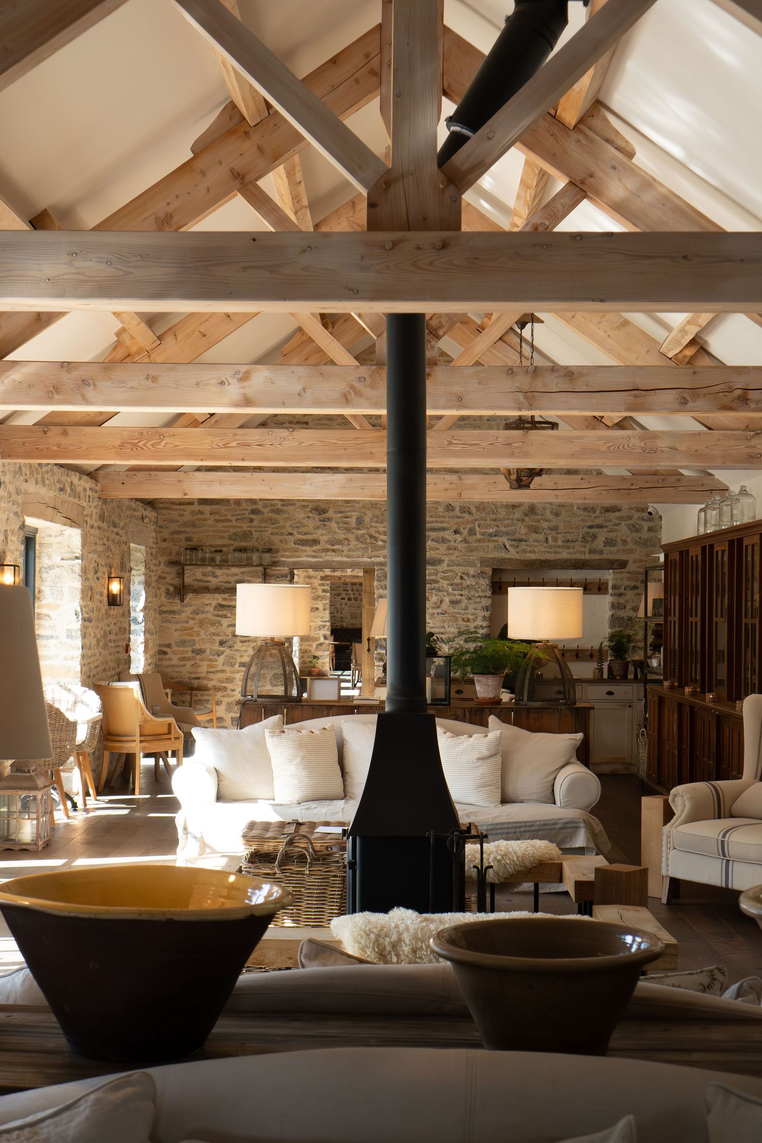 Rustic barn interior with exposed wooden beams, stone walls, and central black fireplace.