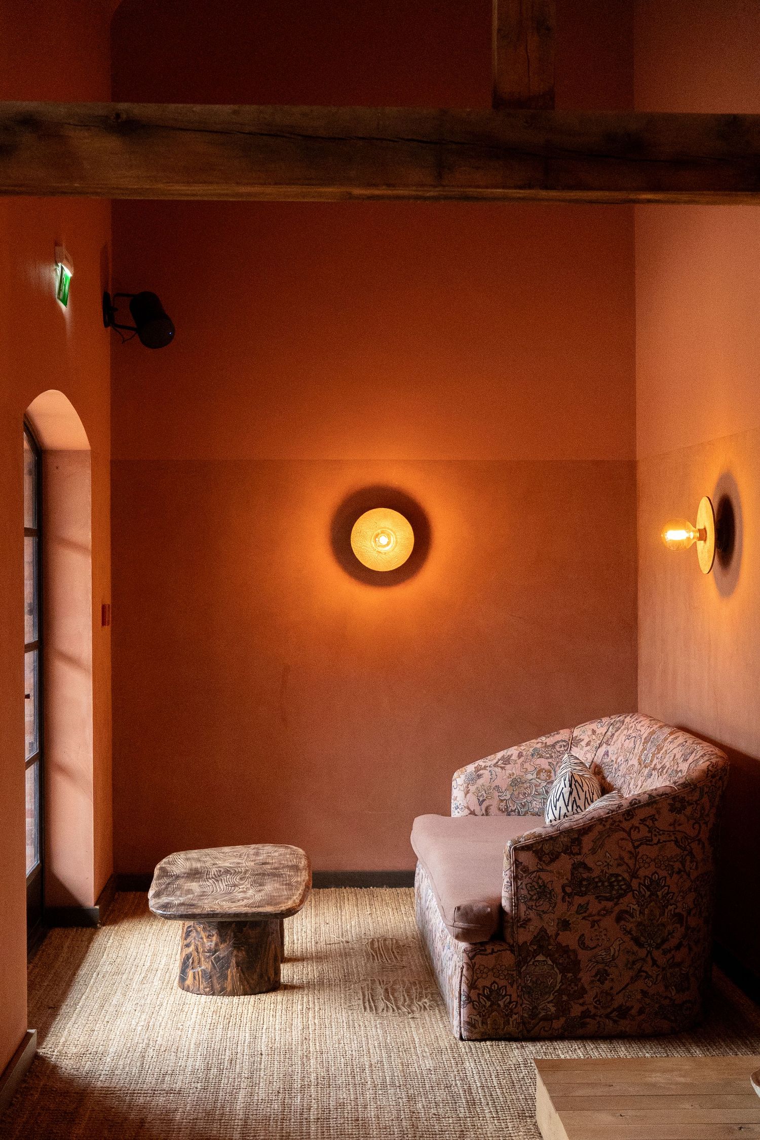Warm terracotta plastered room with floral upholstered sofa, stone side table, and glowing circular wall sconces.
