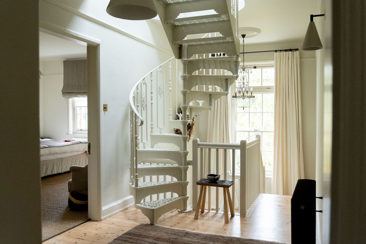 Elegant white spiral staircase in neutral-toned hallway with chandelier, wood floors and sheer curtains.